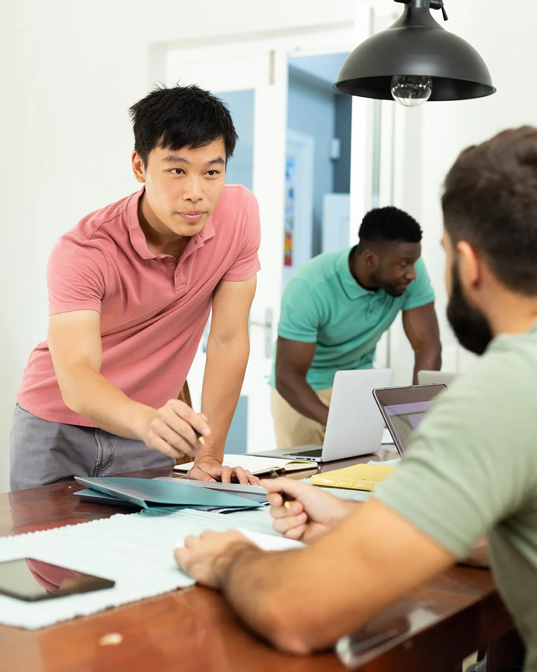 Three men working collaboratively in a bright office, one standing and pointing at documents on the table, two sitting with laptops, engaged in discussion.