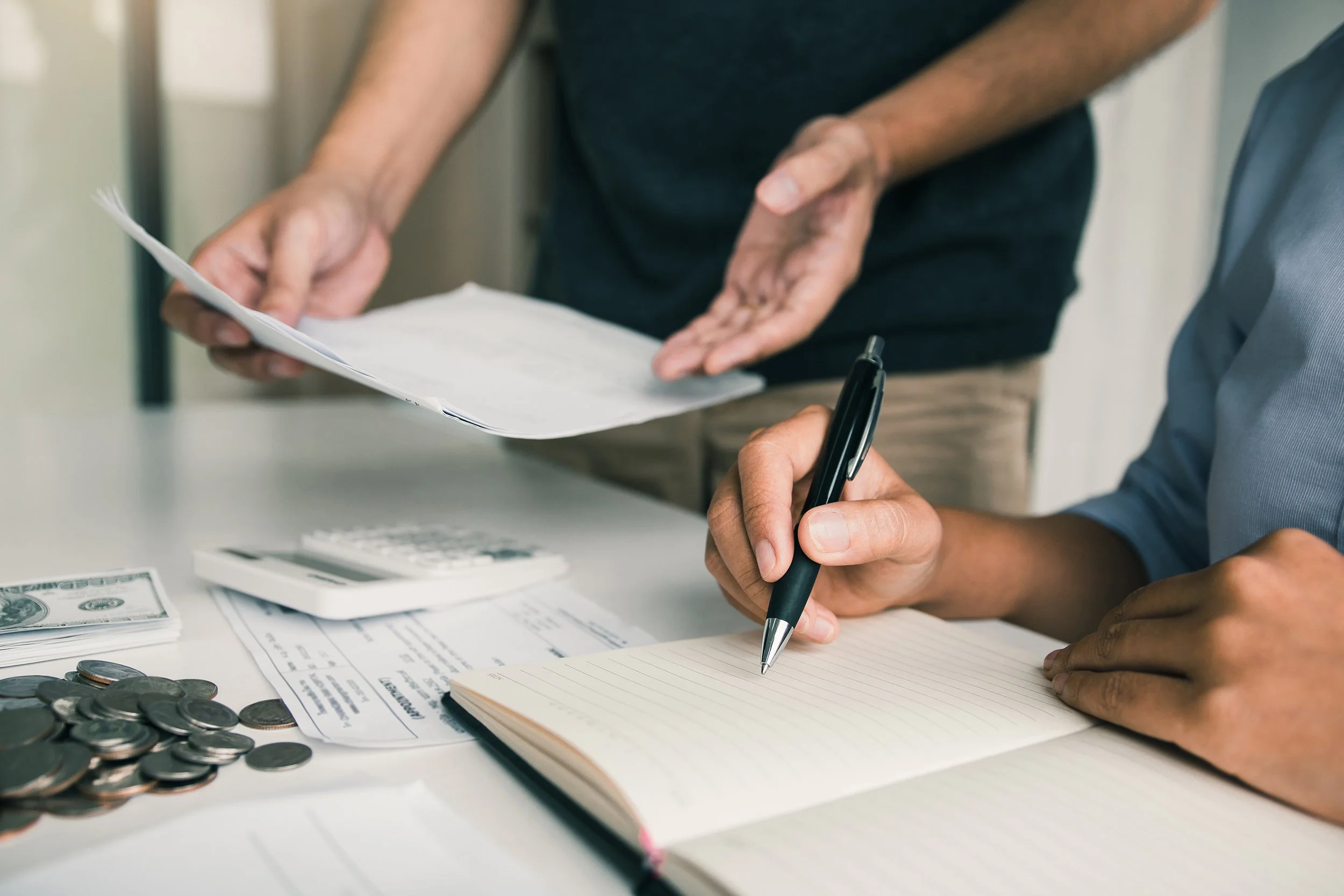 Two people counting money and taking notes, with a calculator, coins, and bills on the table.