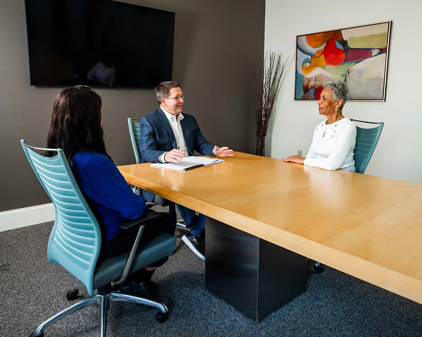 Three people having a business meeting in a conference room with a large wooden table, a flat-screen TV, modern teal office chairs, a colorful abstract painting on the wall, and a tall decorative plant.