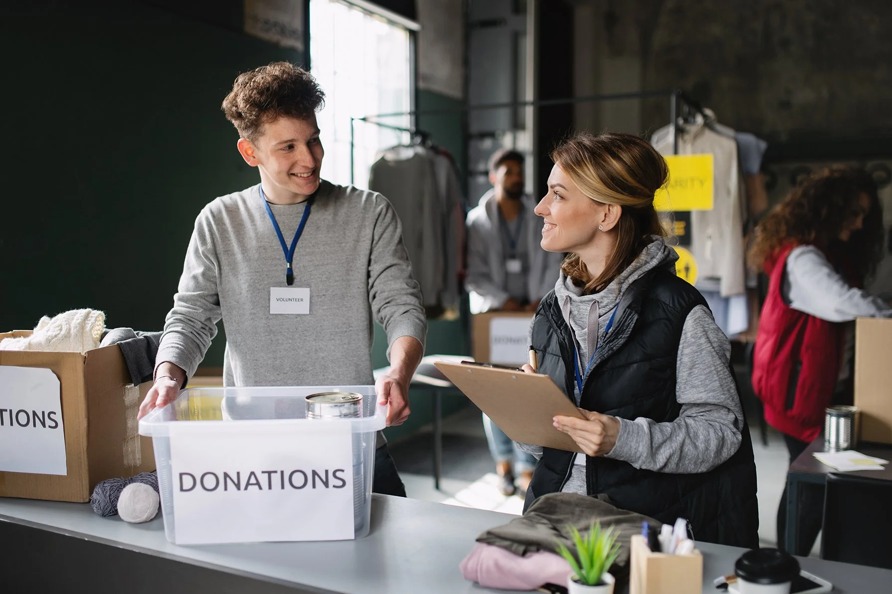 A young male volunteer and a woman are smiling and talking at a donation center. The volunteer is wearing a gray shirt with a name tag, and the woman is holding a clipboard. There are donation boxes and a container labeled 'DONATIONS' on the table. Other volunteers are working in the background.