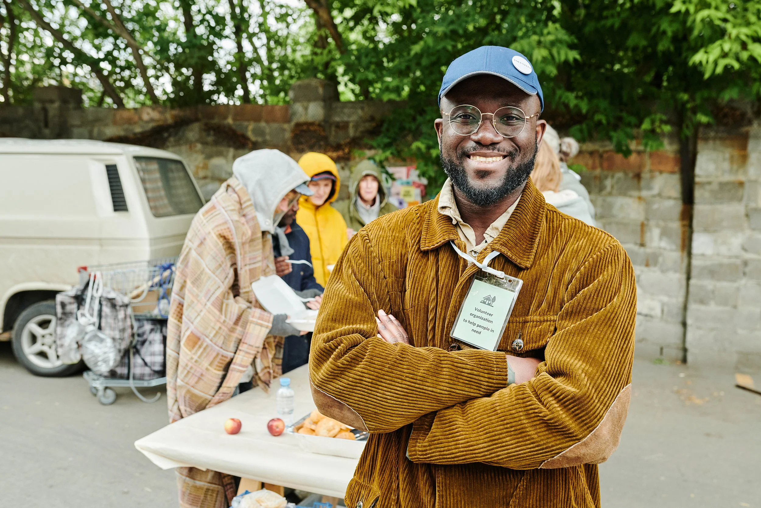 A smiling man with glasses and a blue cap standing with his arms crossed, wearing a brown corduroy jacket and a volunteer badge, outdoors with a table of food and several people in jackets in the background.