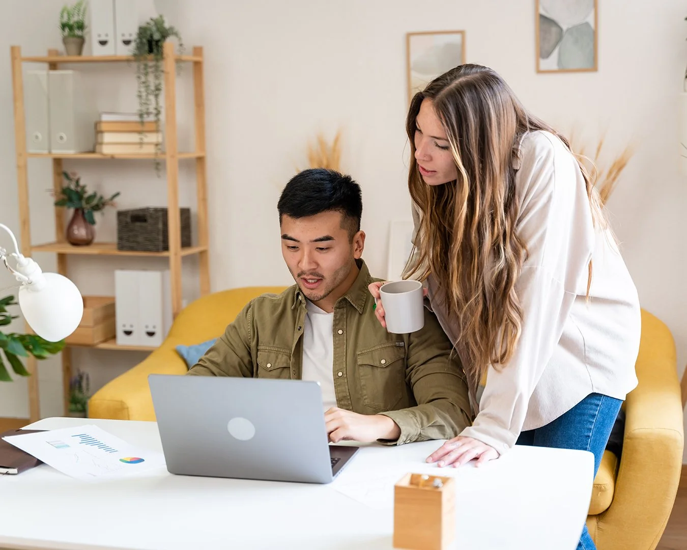Two people working together at a desk with a laptop and papers in a cozy, well-decorated room. One person is seated and typing on the laptop, while the other is leaning over, holding a coffee mug, and looking at the screen.