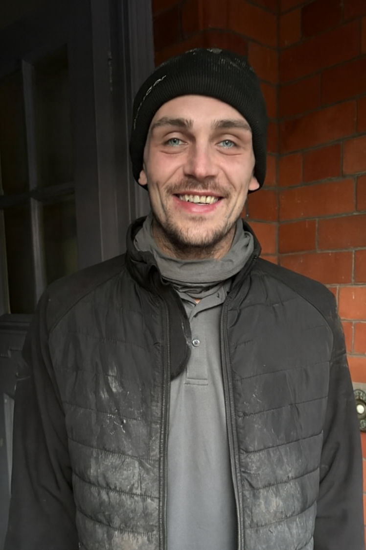 A smiling man wearing a black beanie, gray scarf, gray polo shirt, and black jacket standing outdoors against a brick wall and window.