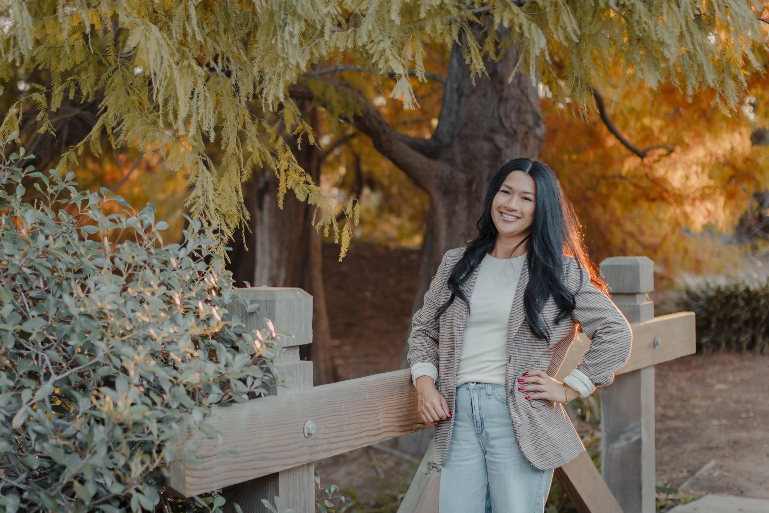 Young woman with long black hair smiling, standing outdoors near a wooden fence during autumn, surrounded by colorful fall foliage.
