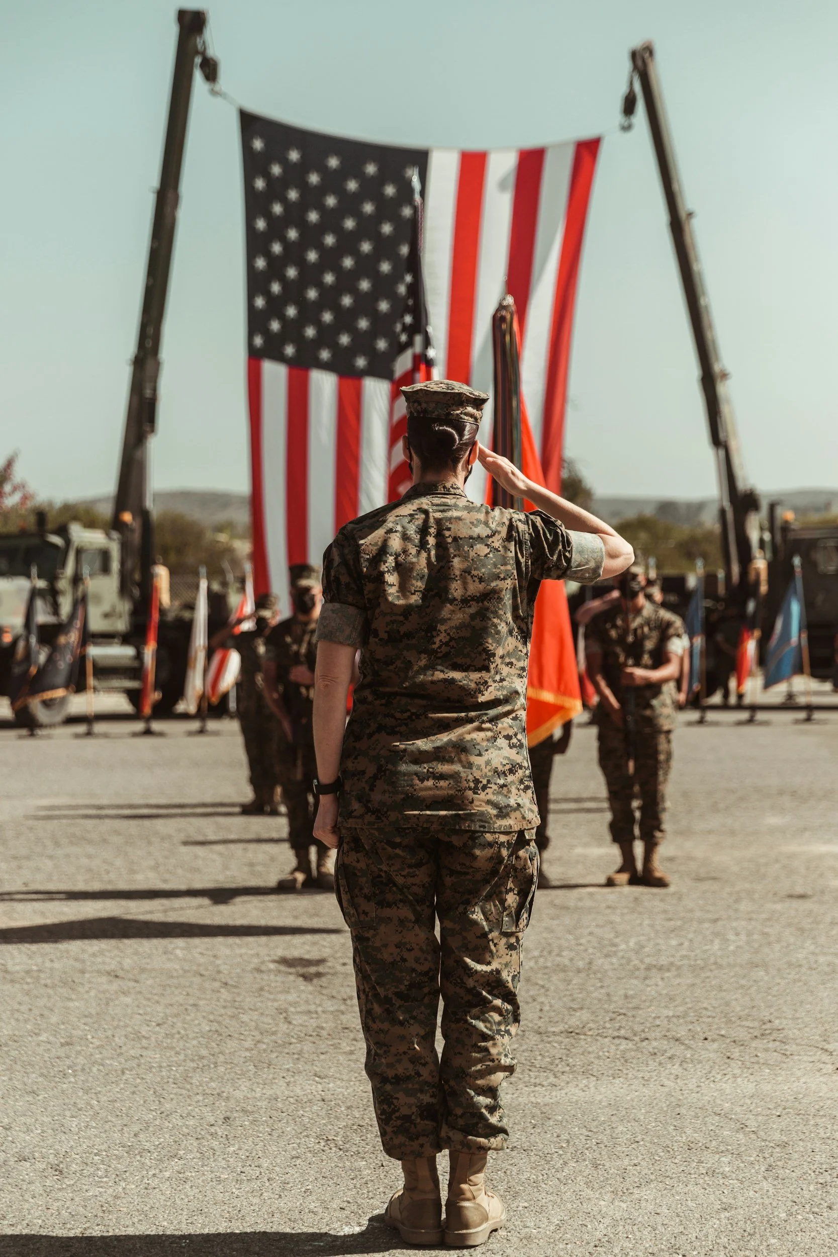 A female soldier in camouflage uniform salutes during a military ceremony with the American flag in the background.