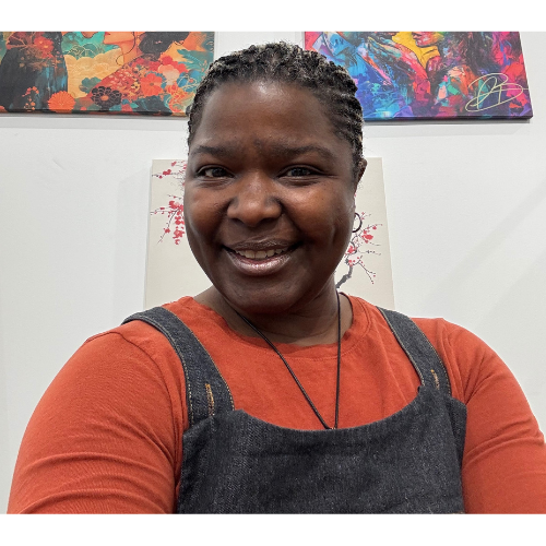 A smiling woman with dark skin, wearing an orange shirt and a black apron, taking a selfie in front of colorful artwork on a white wall.