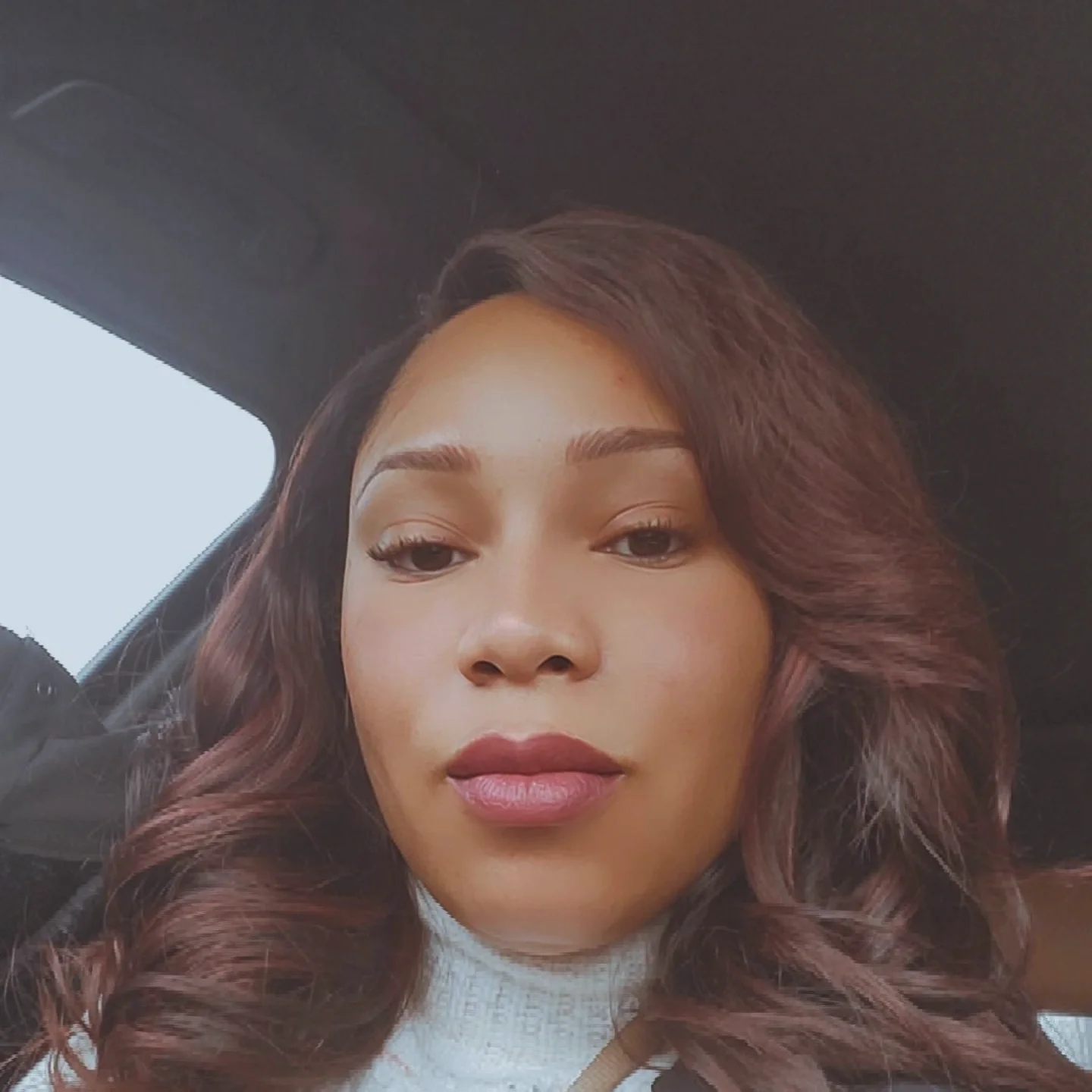 Close-up of a woman with wavy auburn hair, wearing makeup, in a car, with a light-colored turtleneck sweater.