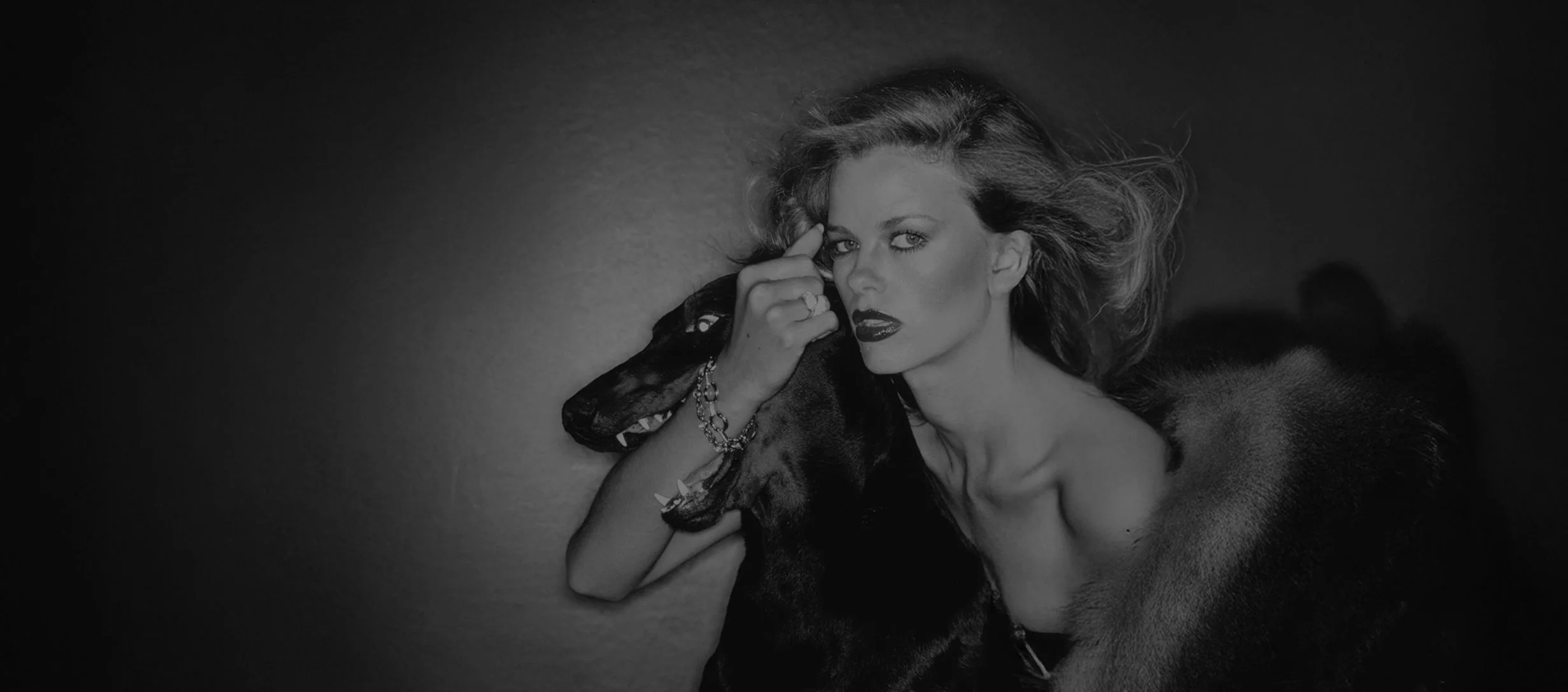 Black and white photo of a woman with wavy hair, wearing dark lipstick, holding a small dog close to her face, and looking directly at the camera.