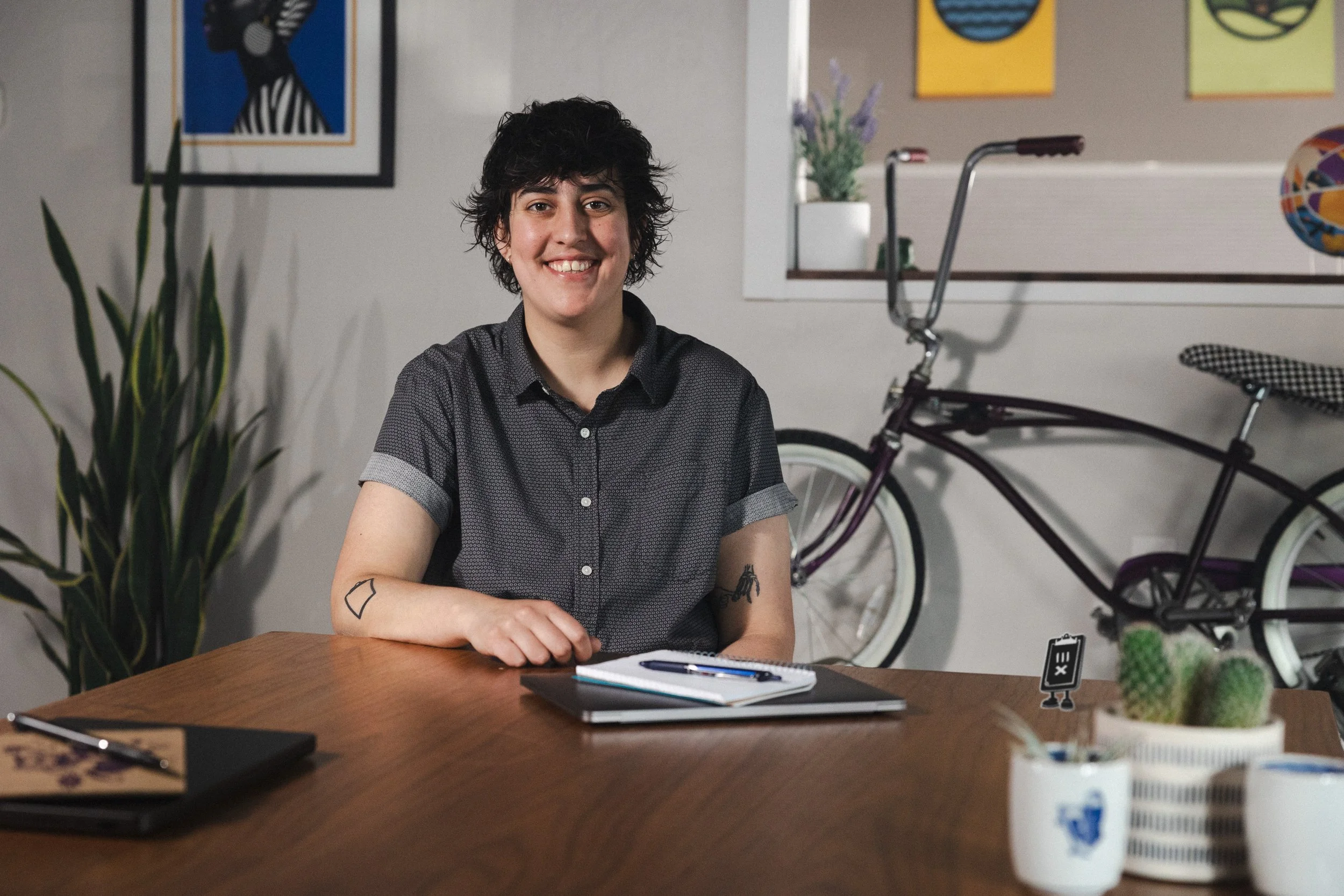 A person with short, curly dark hair, smiling, sitting at a wooden table with notebooks, pens, and a laptop. Indoor setting with plants, framed art, a bicycle mounted on the wall, and decorative items including cacti.