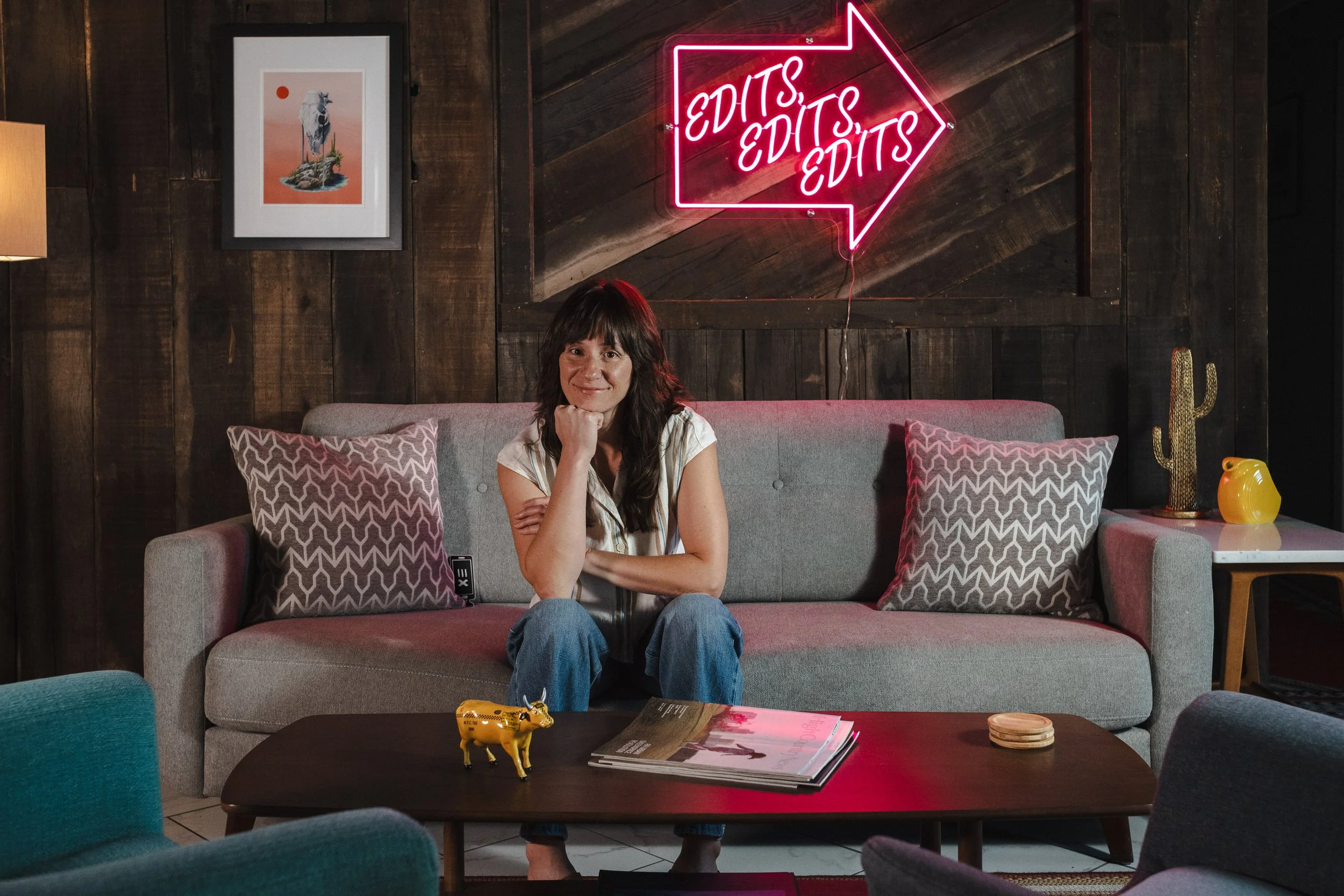 A woman sitting on a gray sofa in a cozy, stylish room with dark wooden walls. Behind her, a neon sign with a pink arrow reads 'EDITS, EDITS, EDITS.' The room has decorative pillows, a small table with magazines, a yellow cow figurine, and a cactus in a pot.