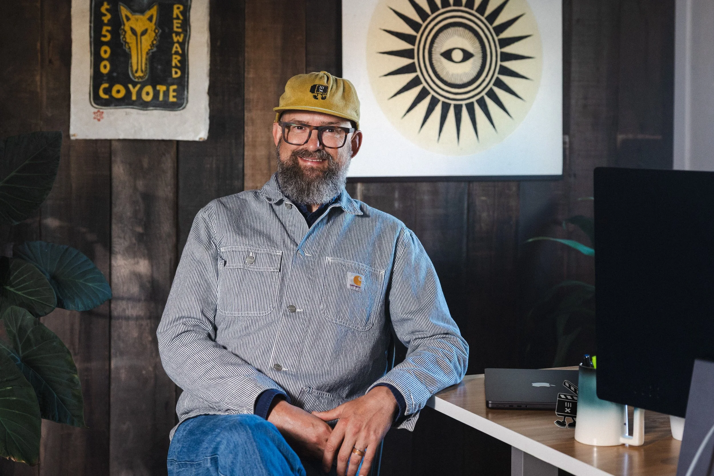 A smiling man with glasses and a beard, wearing a beige cap and a striped gray jacket, sitting at a desk with a closed laptop and a monitor in an office with dark wooden walls and hanging artwork.