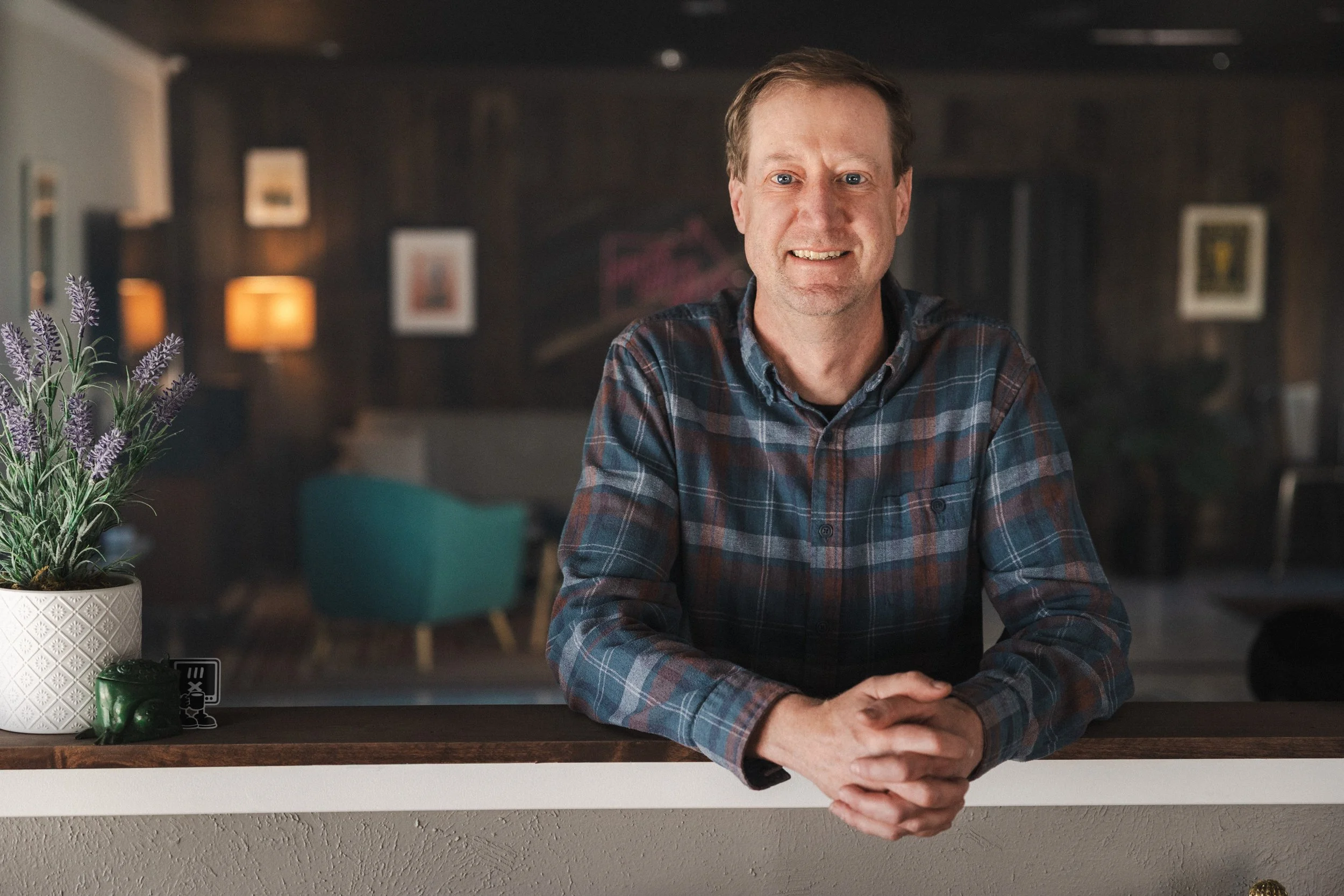 A man in a plaid shirt leaning on a counter in a dimly lit room with framed pictures and lamps in the background.