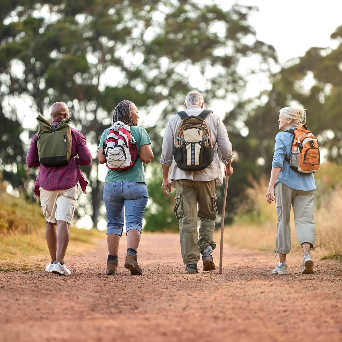 Four people with backpacks hike on a dirt trail in a forested area during daytime.