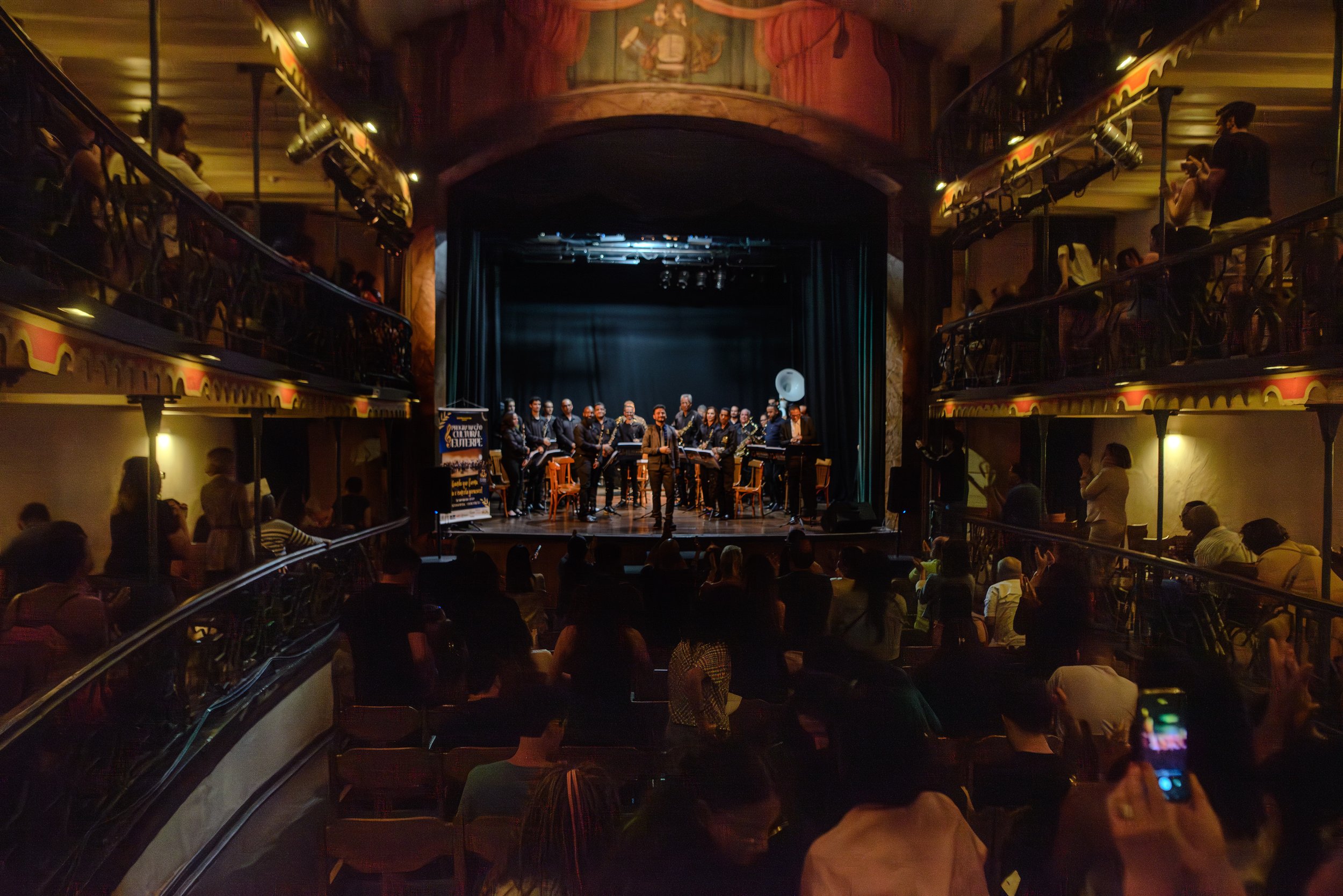 Palco de teatro ou concertos com uma orquestra de músicos de frente para o público, em um espaço com múltiplos níveis e iluminação suave.