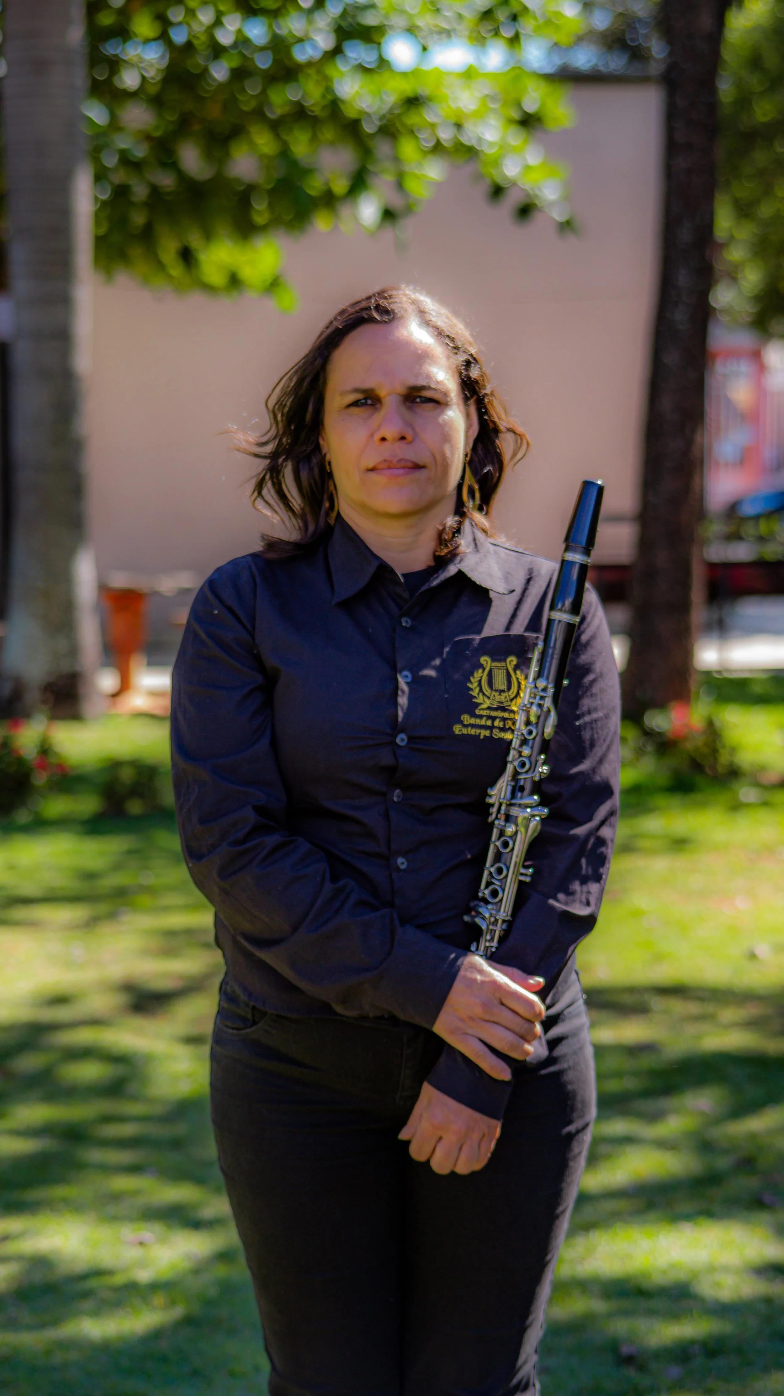 Mulher de cabelos médios encarando a câmera, segurando uma flauta, usando camisa preta com logotipo em um ambiente ao ar livre com árvores e grama.
