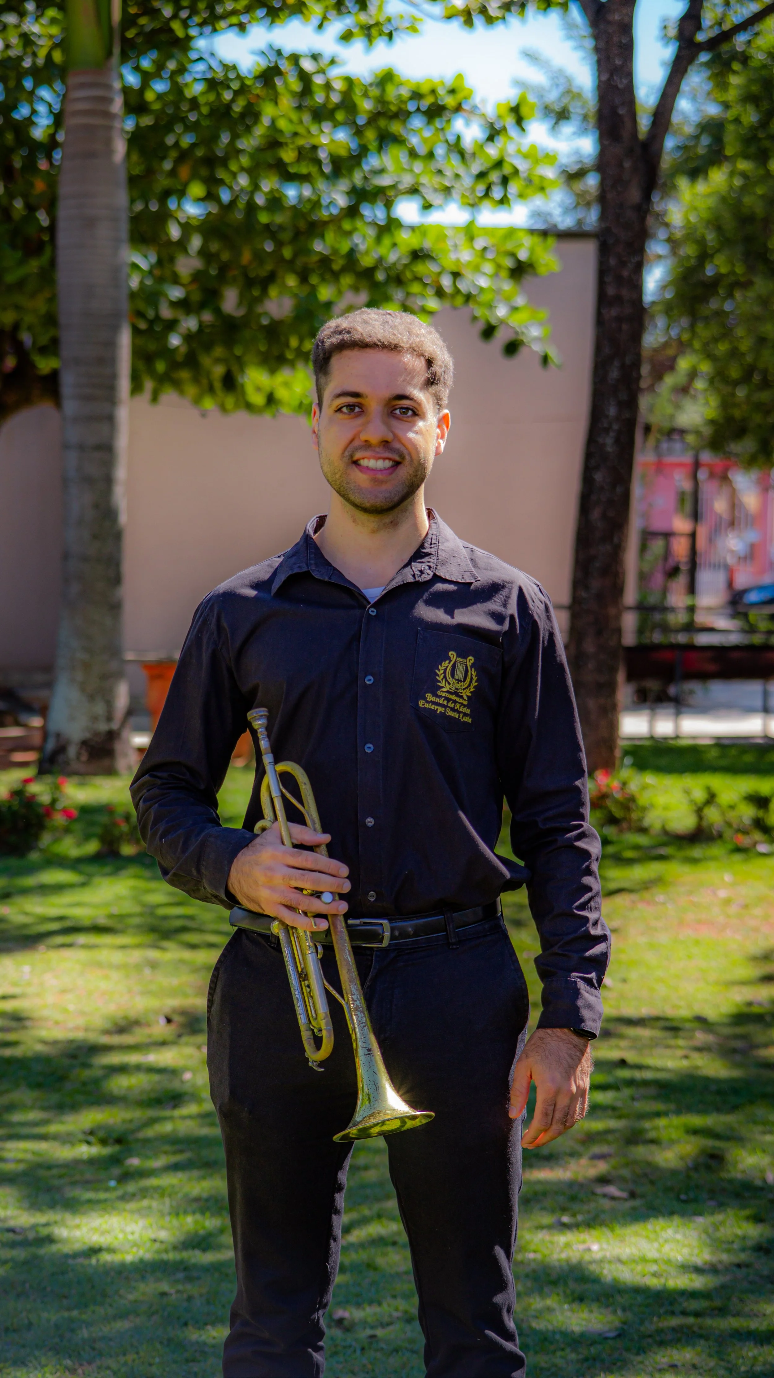 Homem com cabelo encaracolado, barba leve, vestindo camisa preta com símbolo de uma orquestra e texto em português, segurando trompete, em ambiente ao ar livre com árvores e grama.