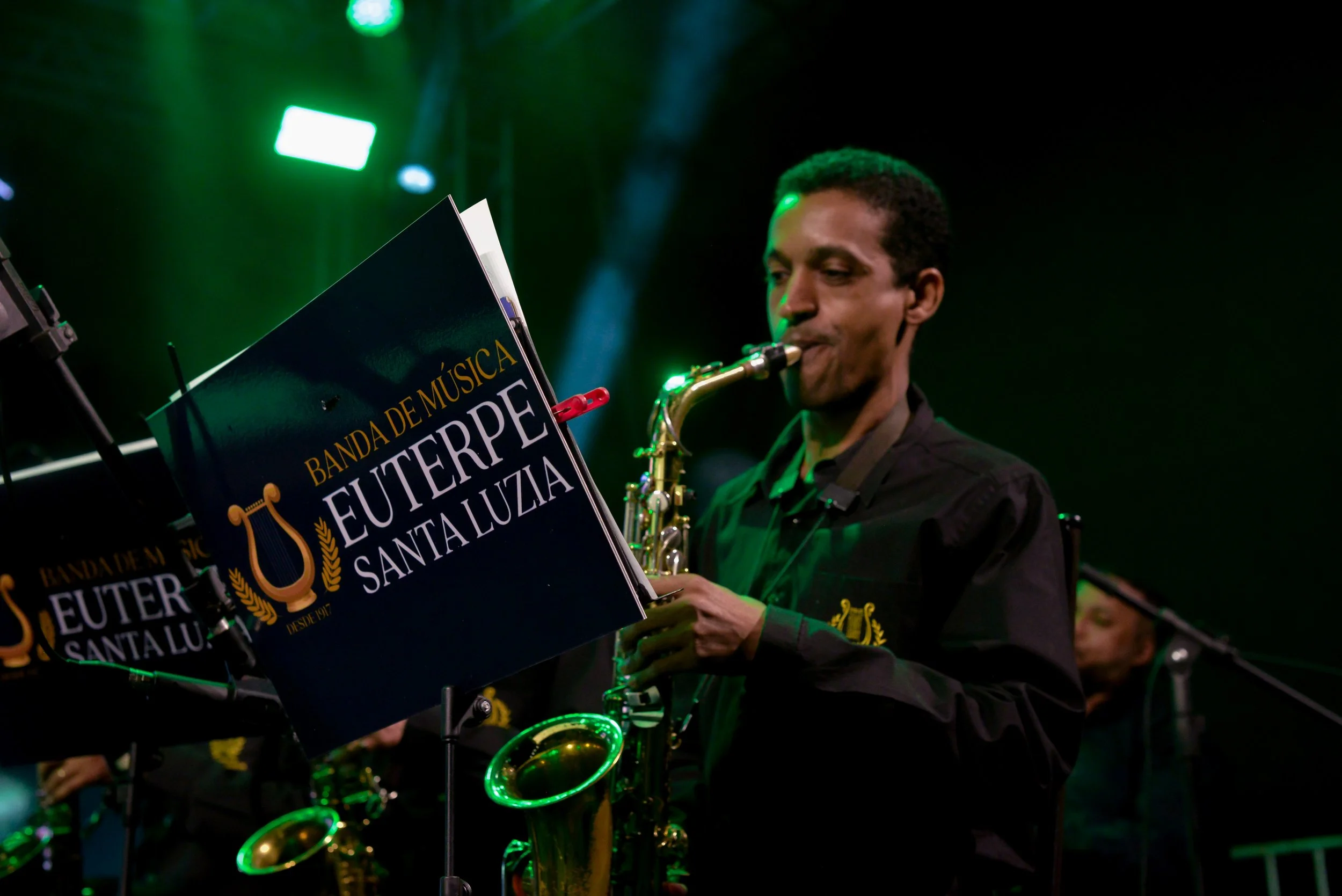 Homem tocando saxofone durante uma apresentação musical, com luzes verdes e fundo escuro, apoiado por uma partitura com o nome "Euterpe Santaluzia".