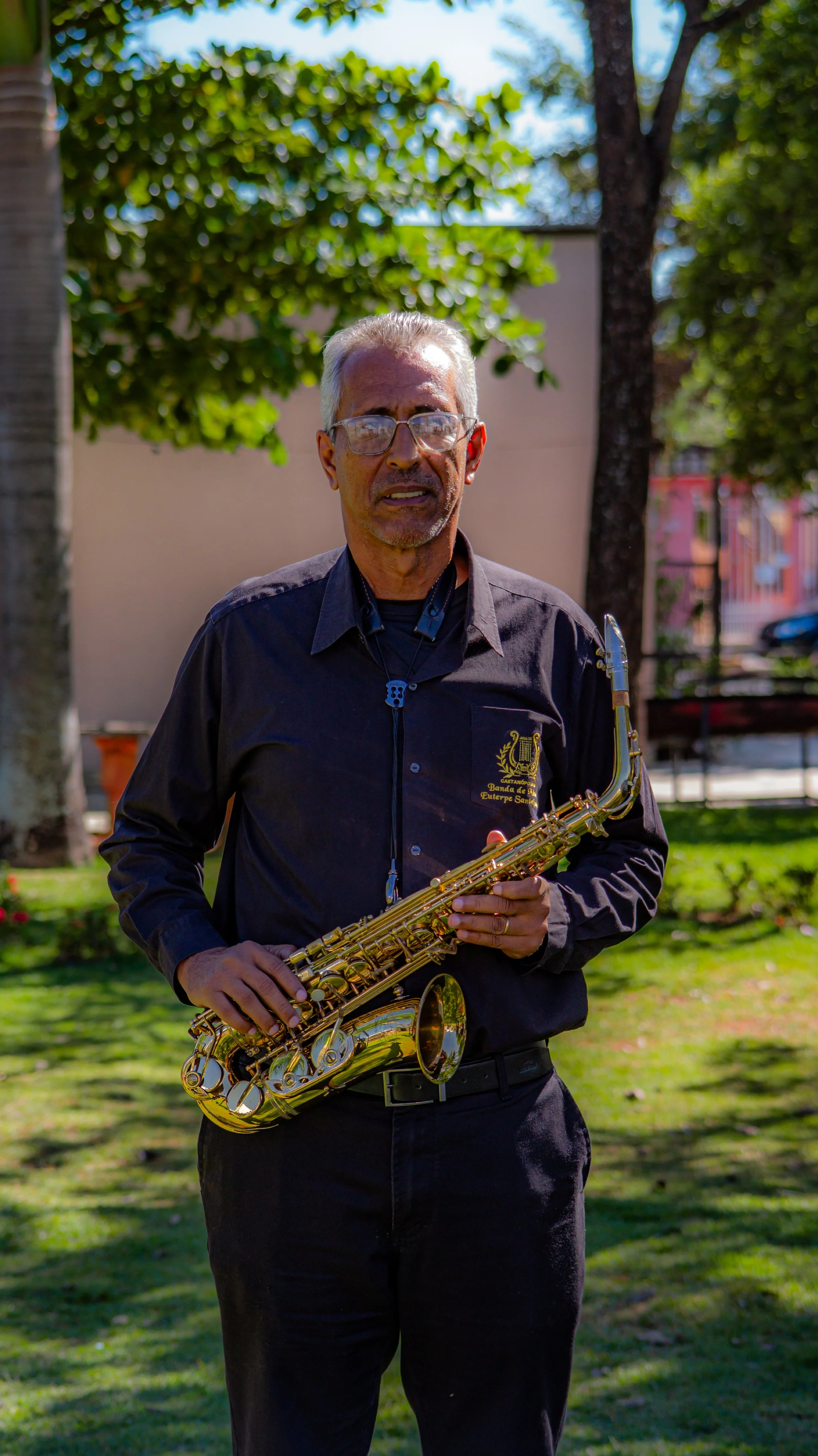 Homem idoso segurando um saxofone em um parque ao ar livre, com árvores e grama, vestindo camisa preta com o símbolo de uma banda