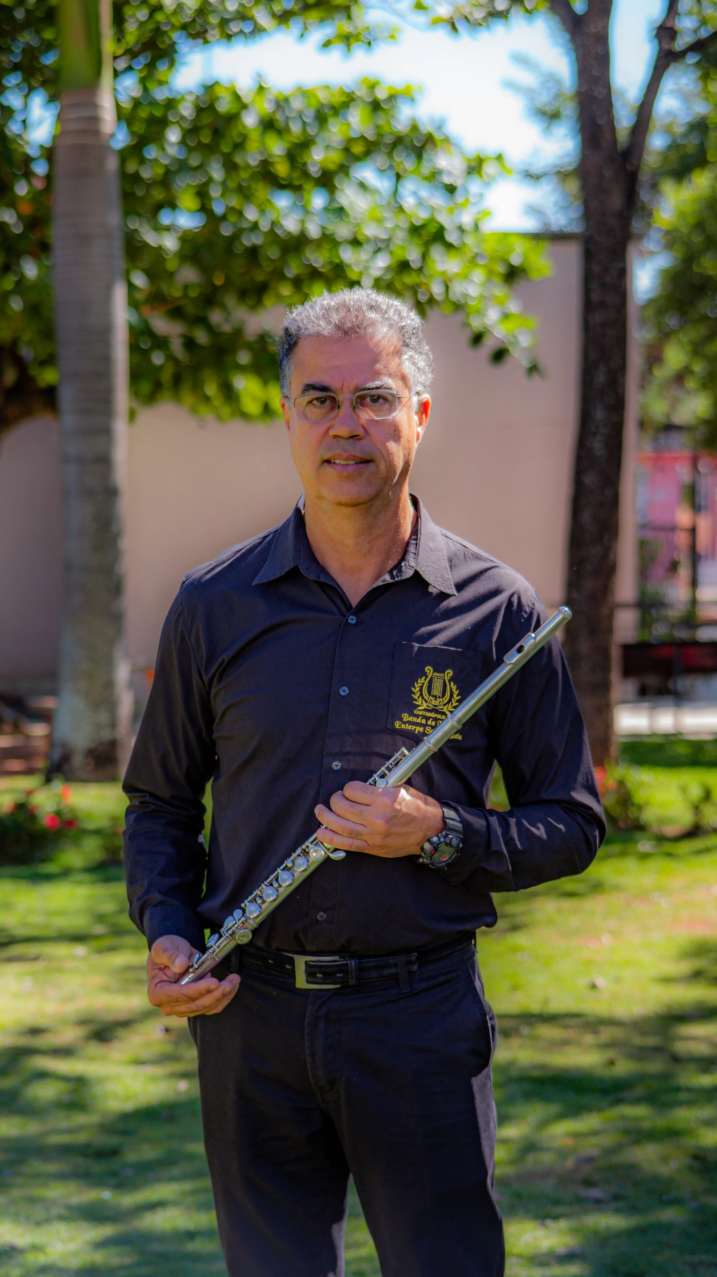 Homem segurando uma flauta transversal posando ao ar livre em um parque com árvores verdes ao fundo.
