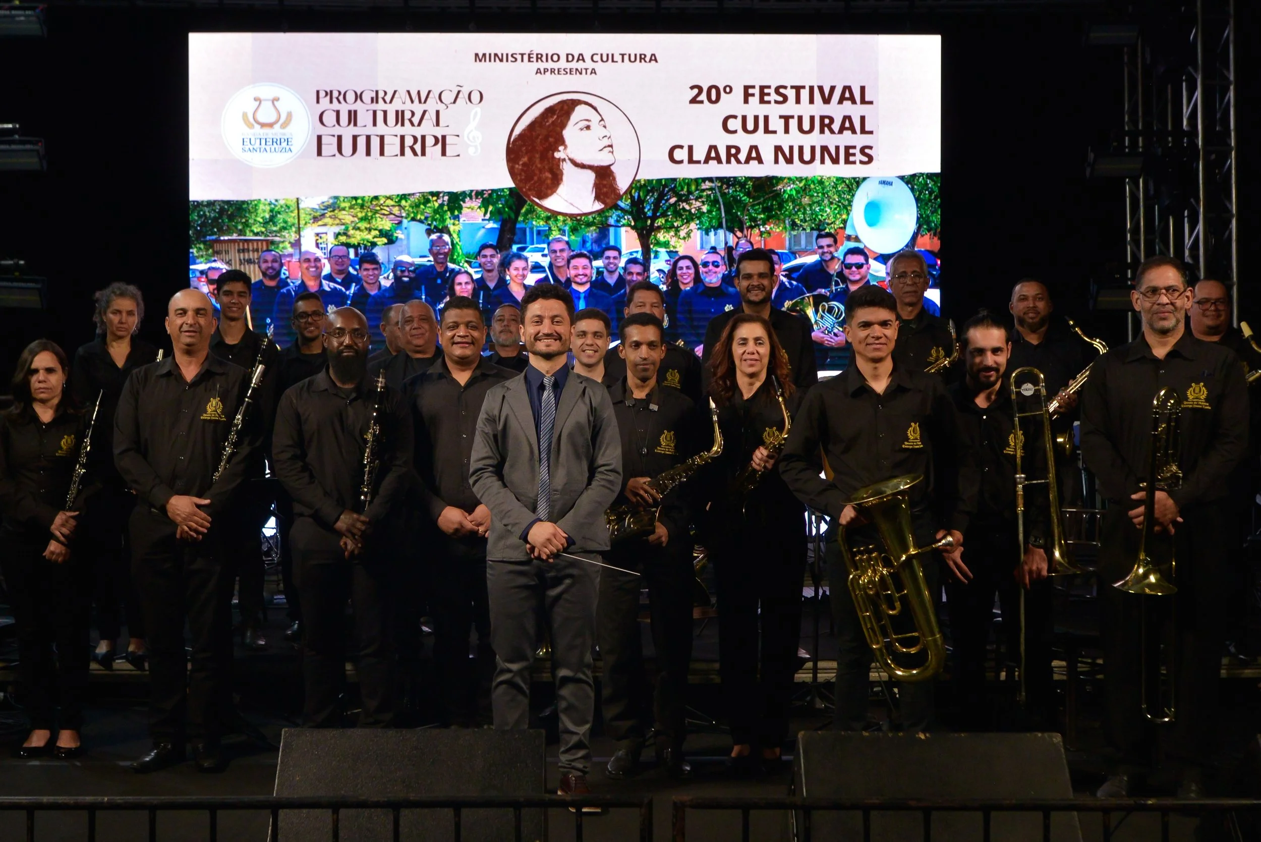 Grupo de músicos no palco com um banner ao fundo que menciona o Festival Cultural Clara Nunes e uma orquestra, com pessoas segurando instrumentos musicais.