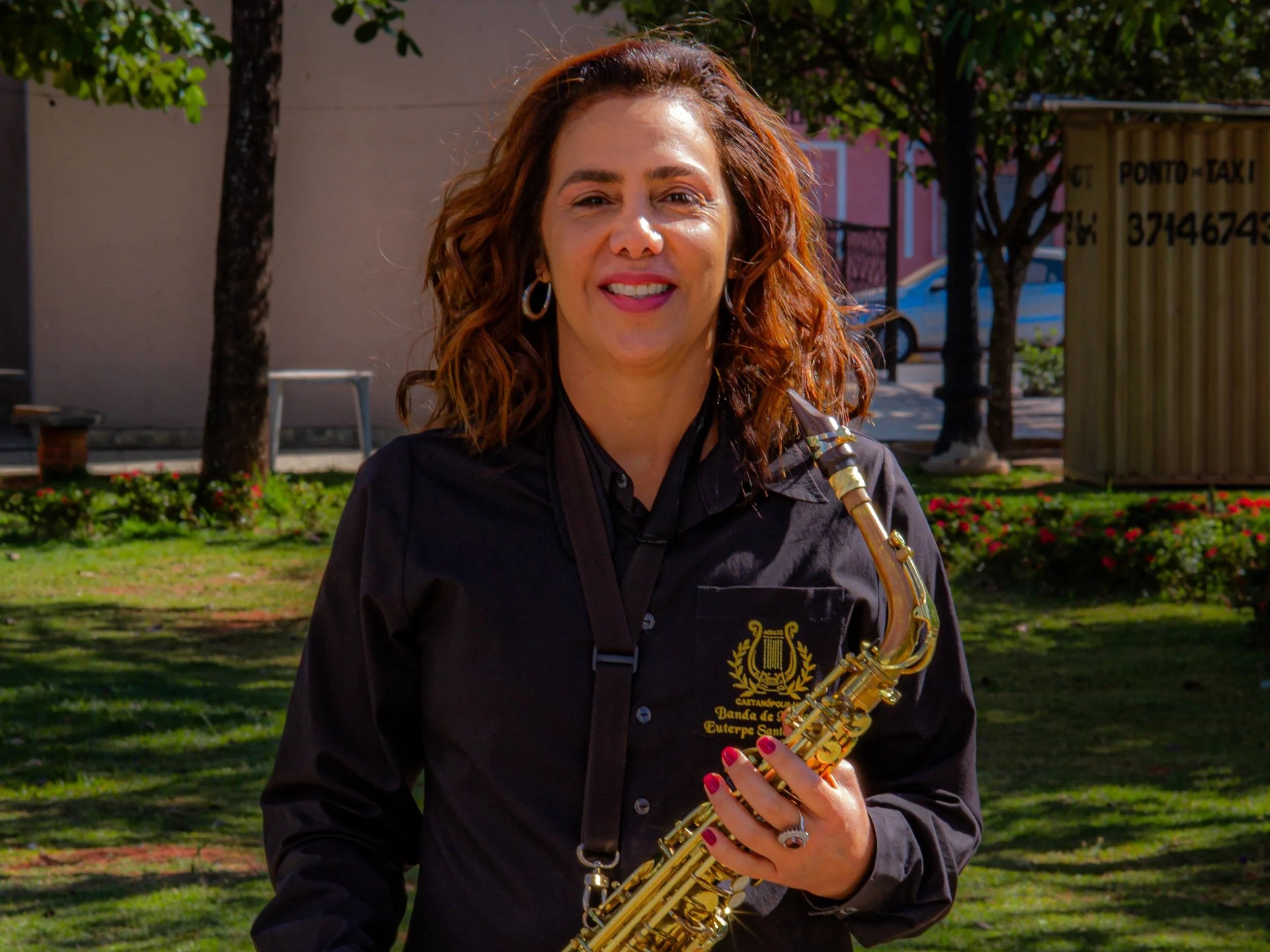 Mulher com cabelo cacheado avermelhado segurando um saxofone dourado ao ar livre, sorrindo, com árvores e céu azul ao fundo.