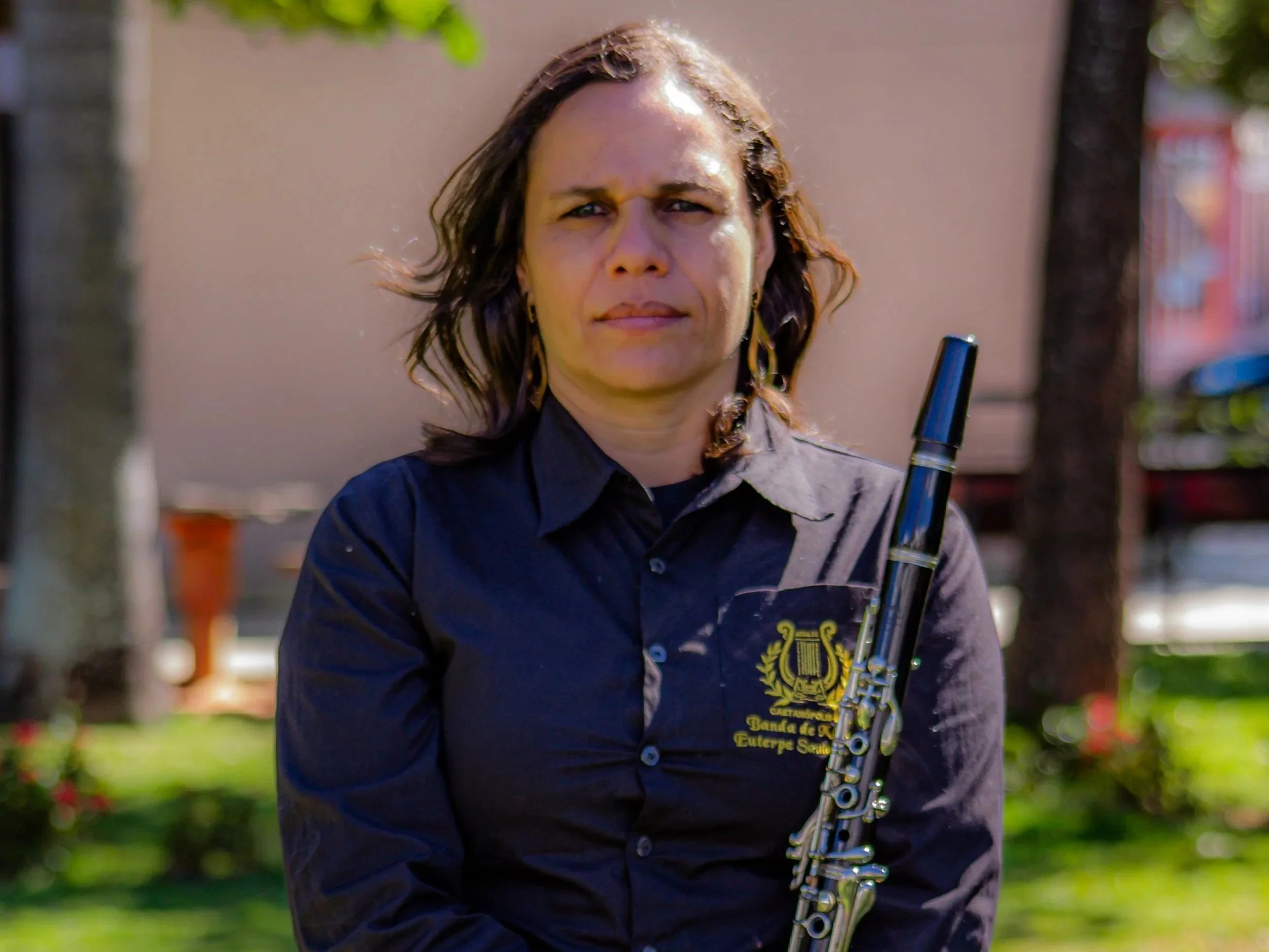 Mulher segurando uma flauta, vestindo uma camisa escura com o logo de uma banda de música, em um ambiente ao ar livre, com árvores e edifícios ao fundo.