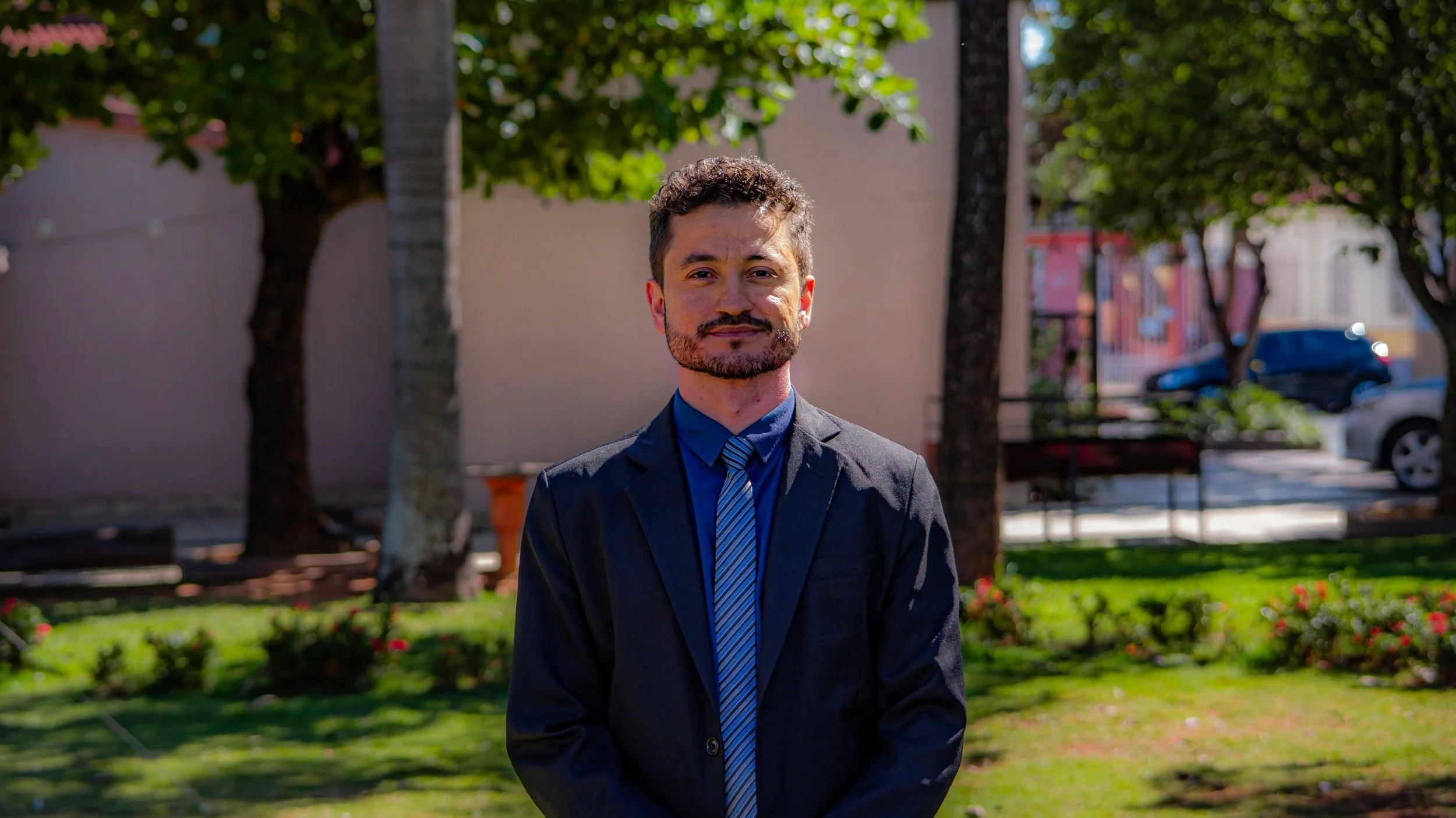 Homem sorridente usando terno escuro, camisa azul e gravata, posando em um parque com árvores e arbustos ao fundo.