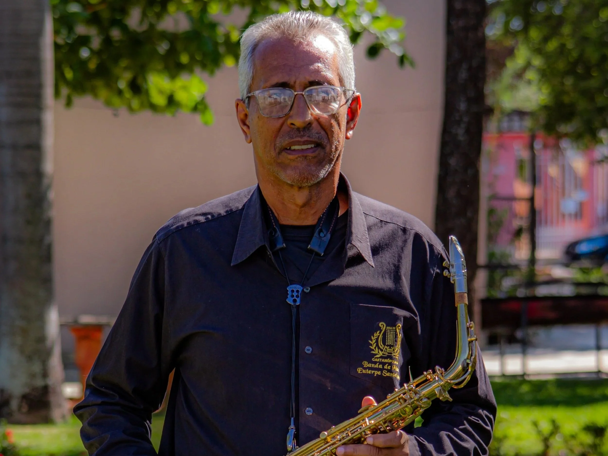 Homem idoso com cabelos brancos, óculos transparentes, vestindo camisa preta com logotipo de uma banda de música, segurando um saxofone dourado, em um parque com árvores e edifícios ao fundo.