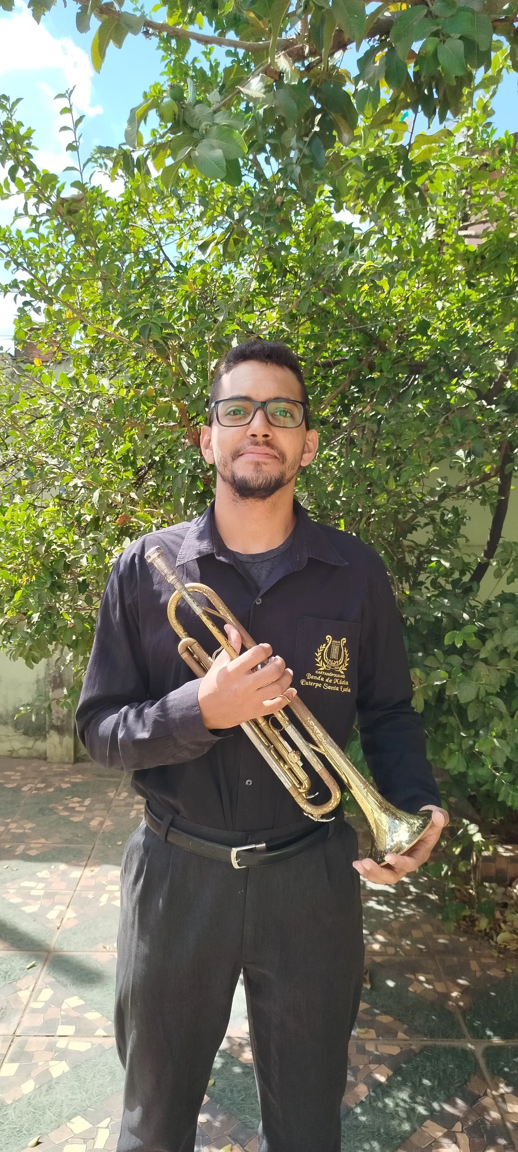 Homem jovem com barba e óculos segurando um trompete, vestindo camisa preta com logotipo na frente e chapéu de aba larga, ao ar livre em um jardim com árvores verdes.