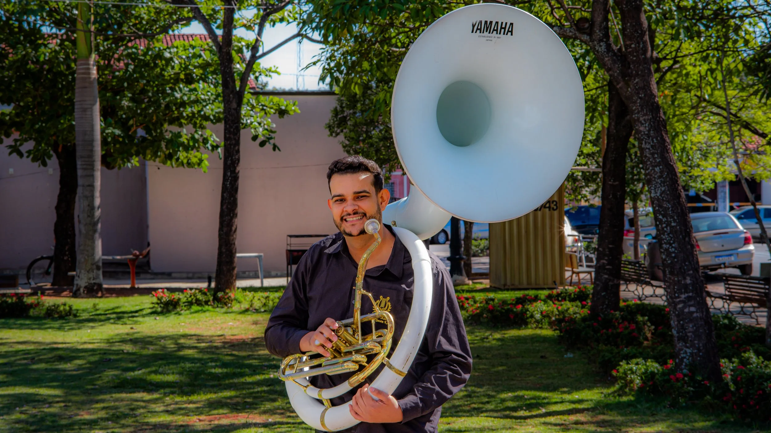 Jovem tocando instrumento musical sousafone ao ar livre, rodeado de árvores e plantas.