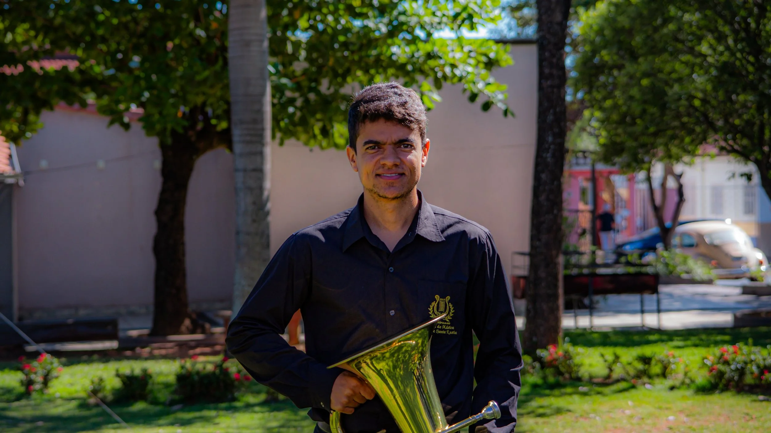 Jovem músico segurando um trombone em um parquinho com árvores, grama e árvores ao fundo.