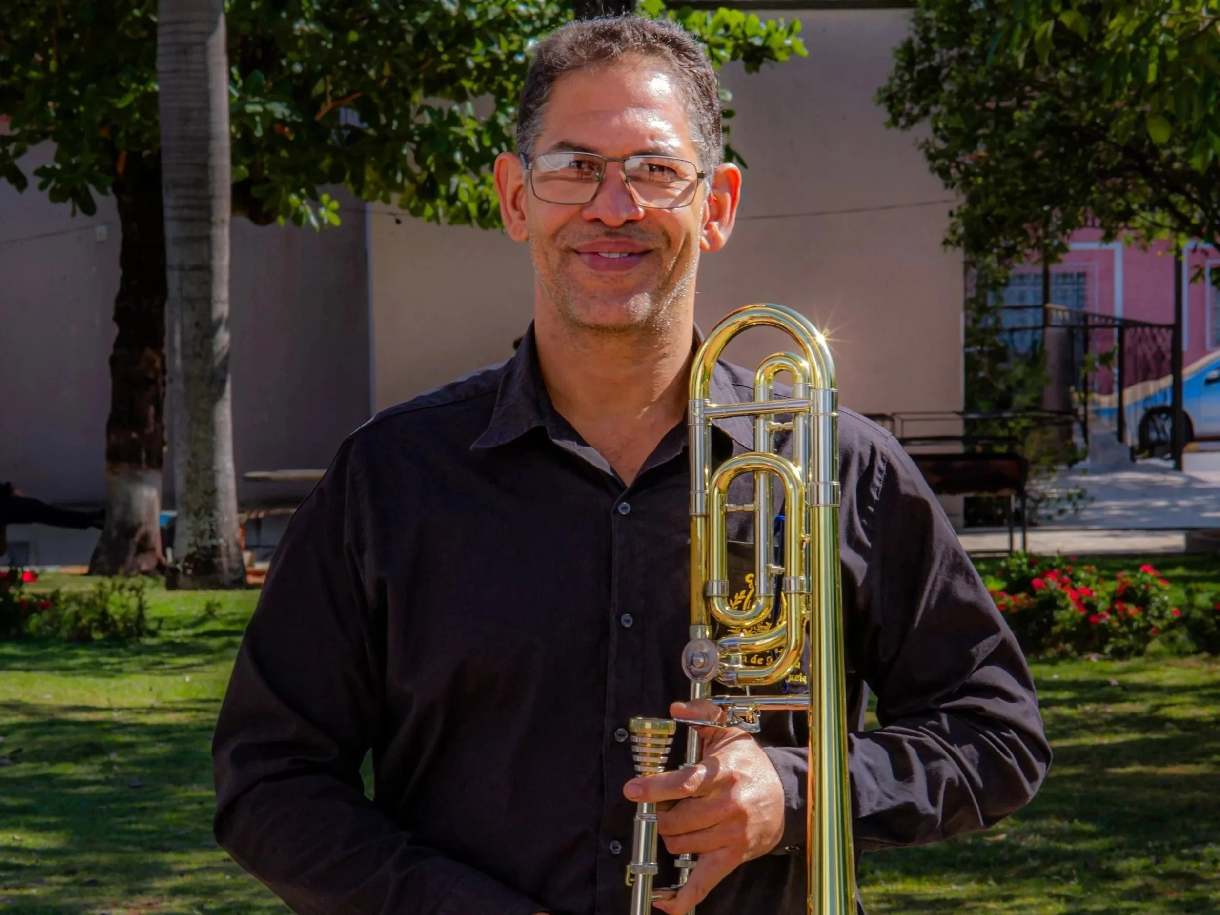 Homem sorridente segurando um trompete de ouro ao ar livre, ao fundo árvores, uma praça e casas coloridas.