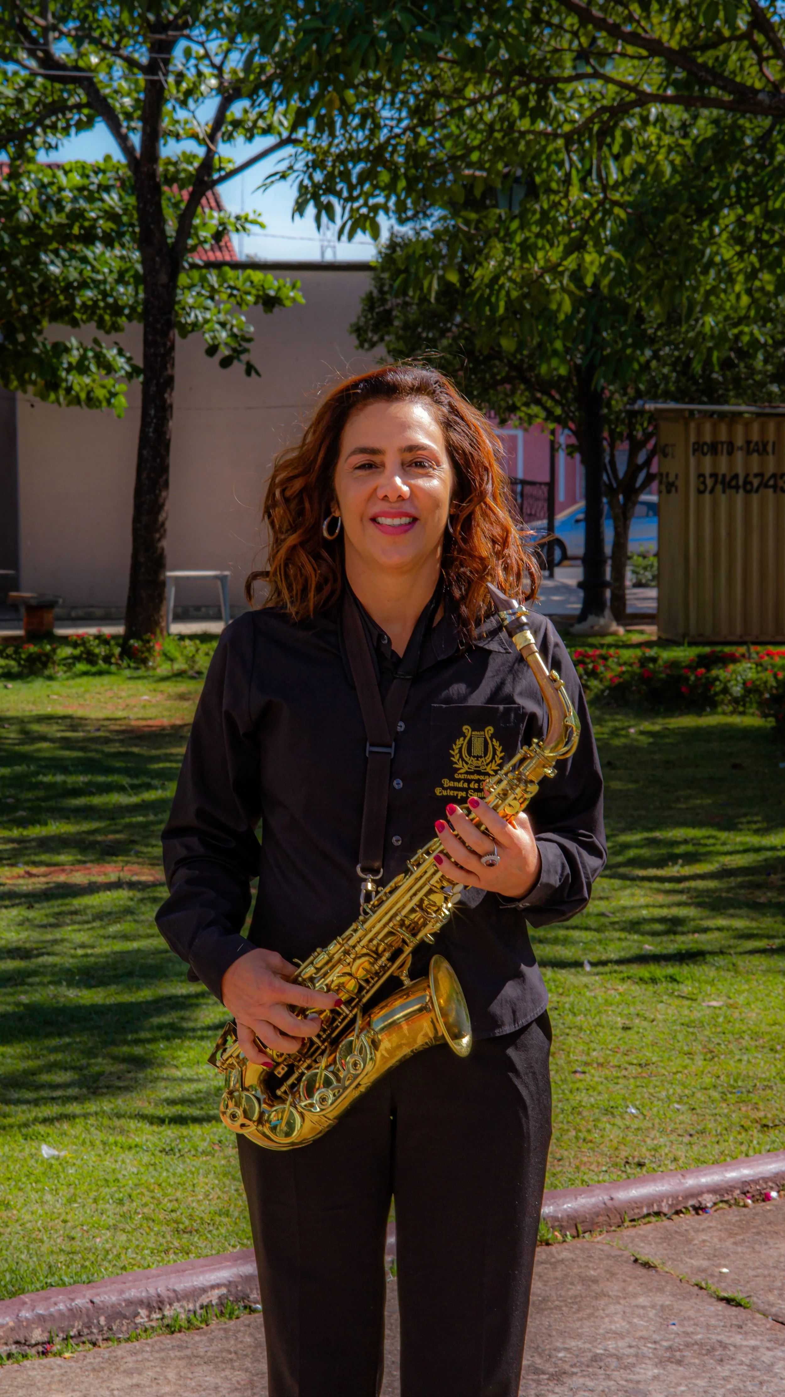 Mulher com cabelo cacheado, vestindo blusa preta com emblema, segurando um saxofone dourado em um parque ao ar livre em um dia ensolarado.