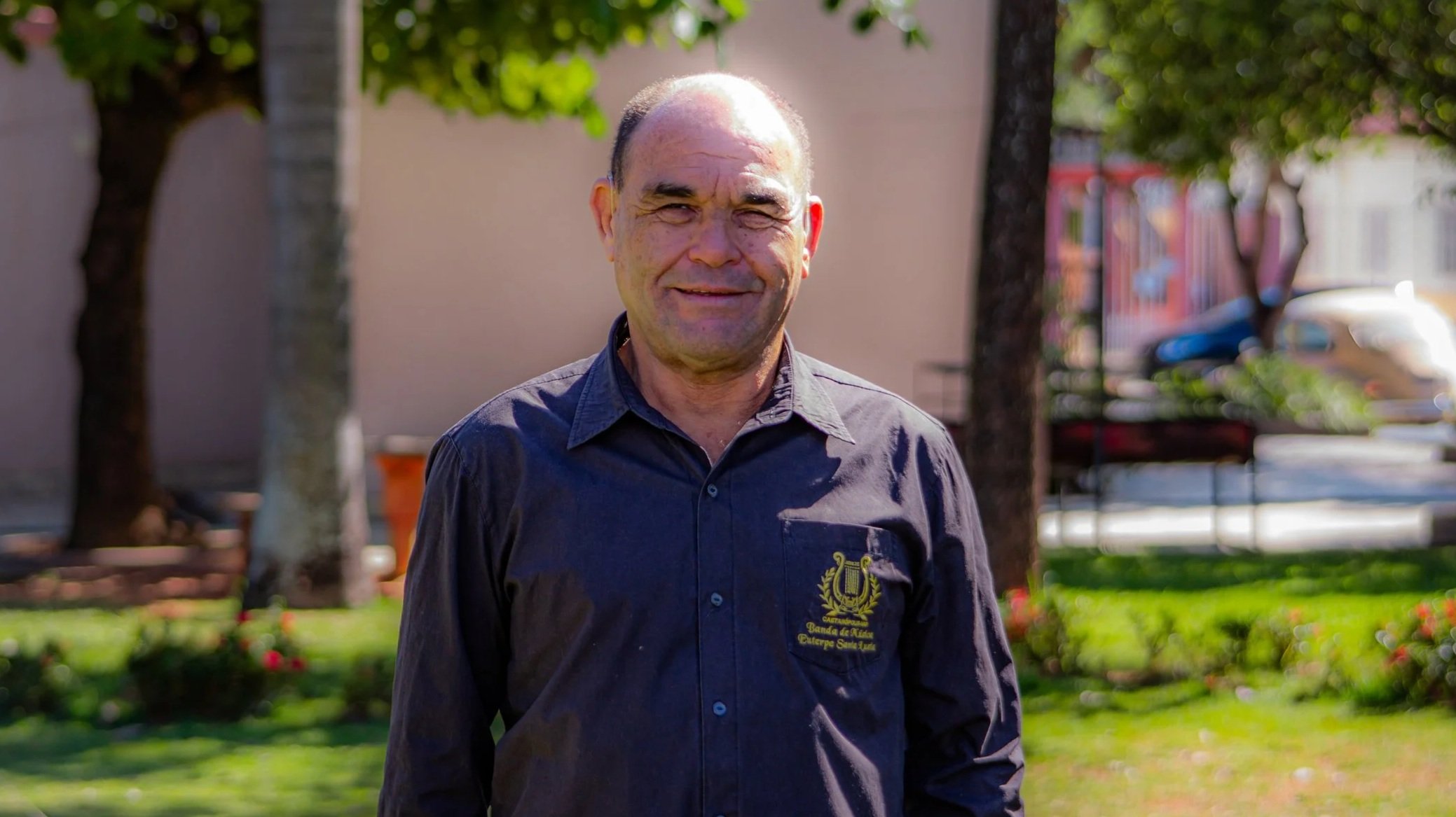 Homem sorridente usando camisa com logotipo em um parque ao ar livre em um dia ensolarado.