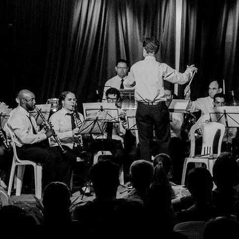 Orquestra de músicos tocando no palco com direção de maestro, em um ambiente com cortinas de fundo, público assistindo.