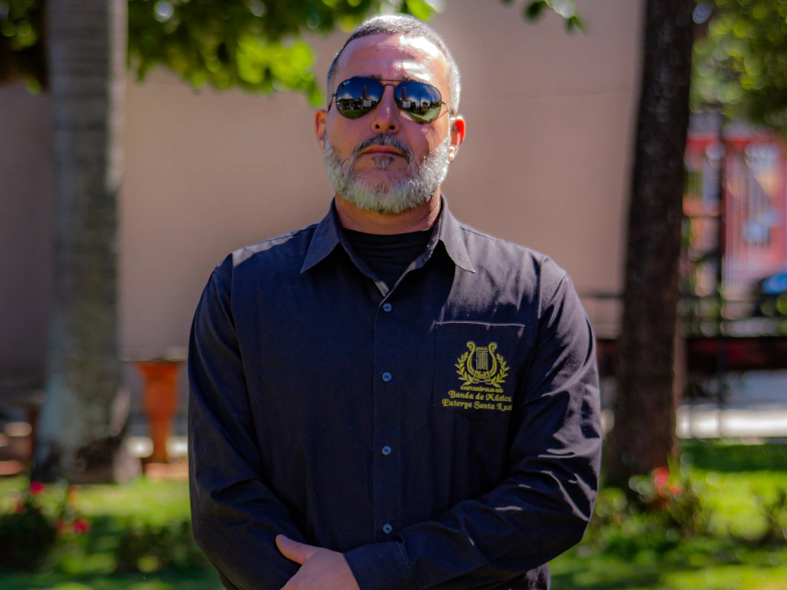 Homem de barba cinza, usando óculos escuros e camisa preta com um emblema em um ambiente ao ar livre com árvores e uma parede ao fundo.