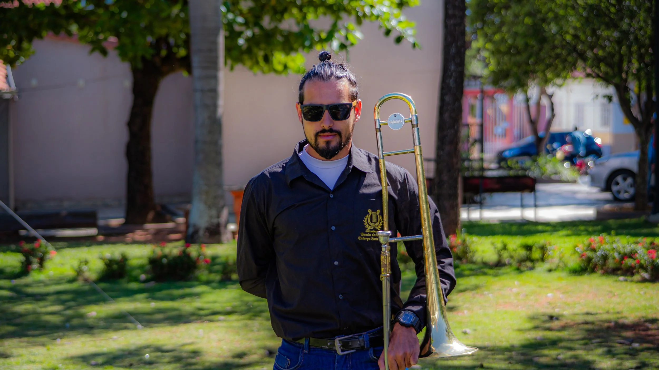 Homem com barba e óculos escuros segurando um trompete em um parque.