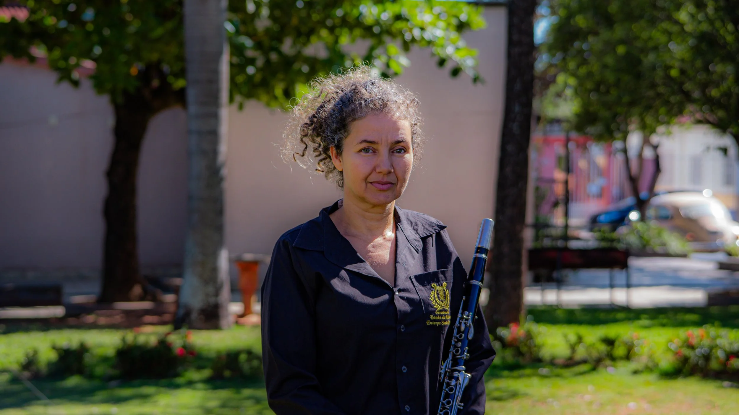 Mulher com cabelo cacheado, vestindo camisa preta com emblema, segurando uma flauta, em um parque com árvores e grama.