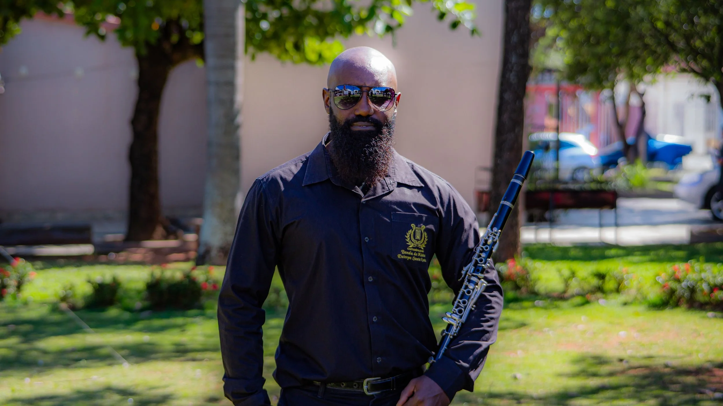 Homem de óculos escuros, barba longa, vestido de camisa preta, segurando uma flauta, em parque com árvores e gramado