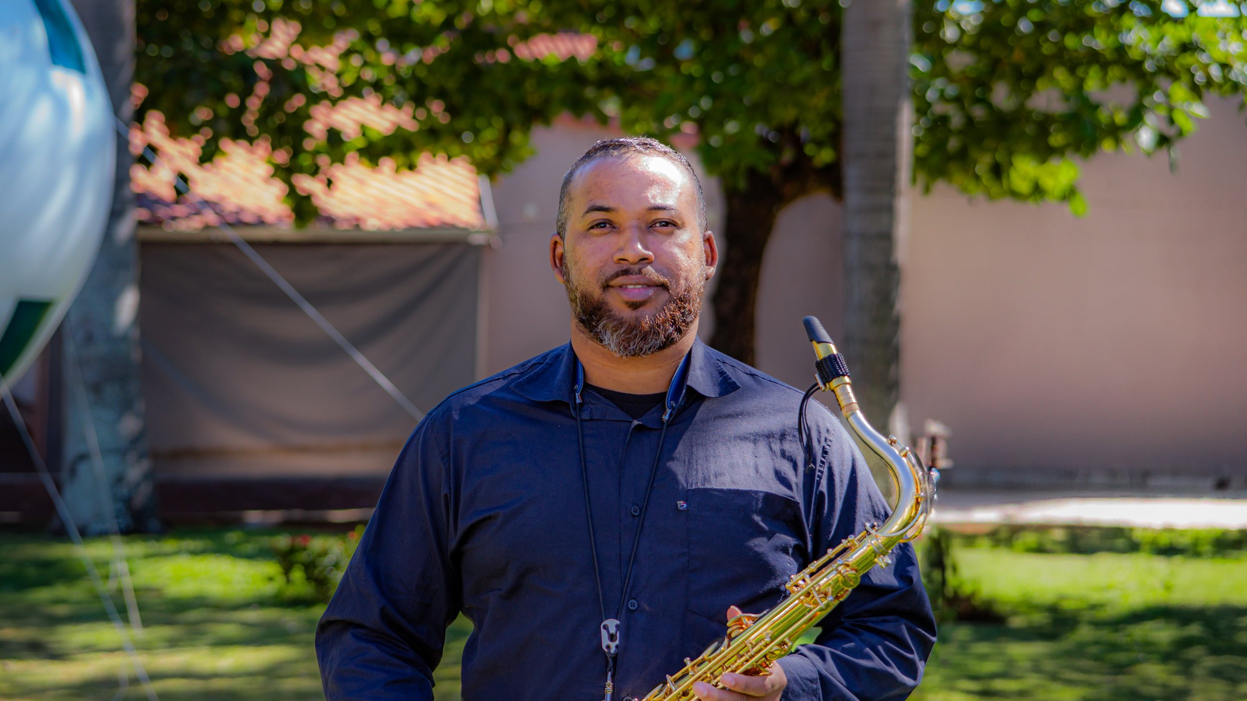 Homem segurando saxofone ao ar livre, com árvores e uma parede ao fundo.