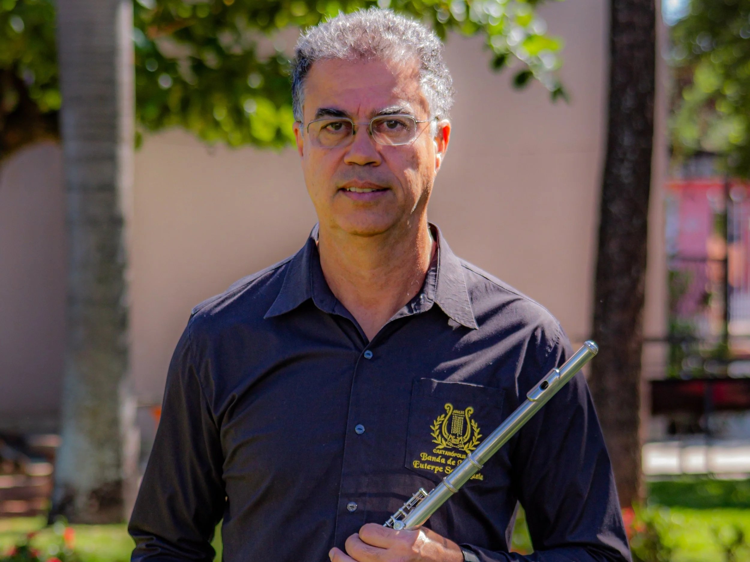 Homem com cabelos grisalhos, óculos e camisa preta com logo de banda de música, segurando uma flauta transversal, em um parque com árvores ao fundo.
