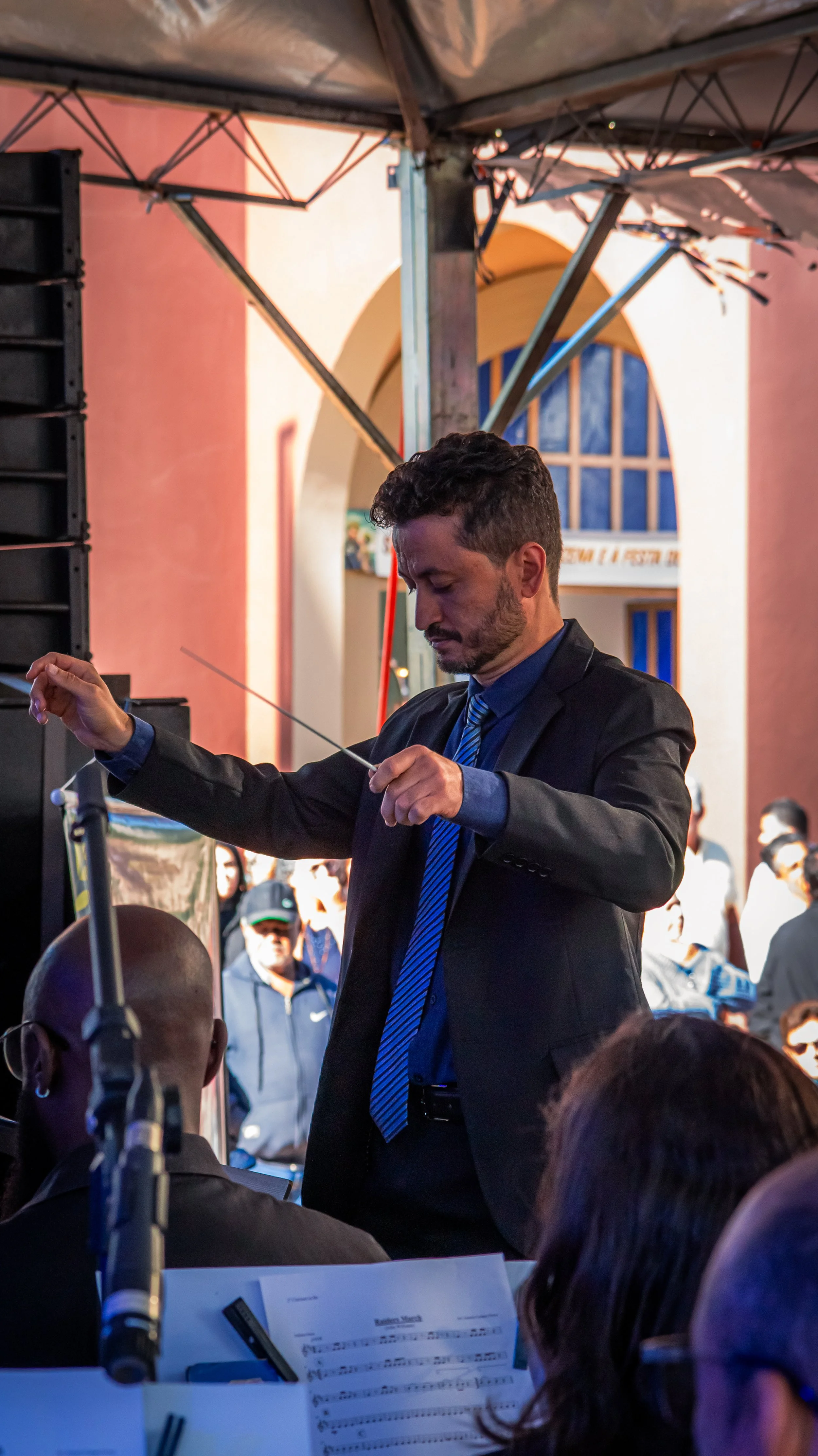 Maestro de orquestra conduz uma apresentação musical em um ambiente interno, com vários espectadores ao fundo.