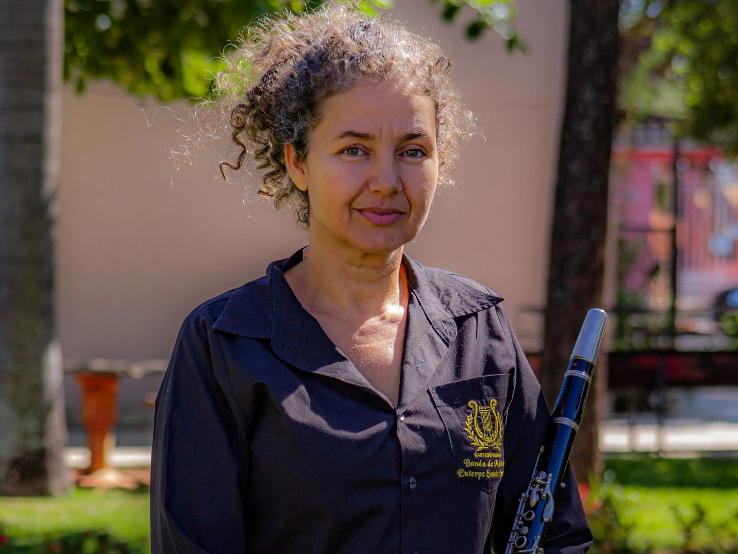 Mulher com cabelo cacheado acinzentado, usando camisa preta com um escudo e palavras em português, segurando uma flauta, ao ar livre com árvores e muro ao fundo.