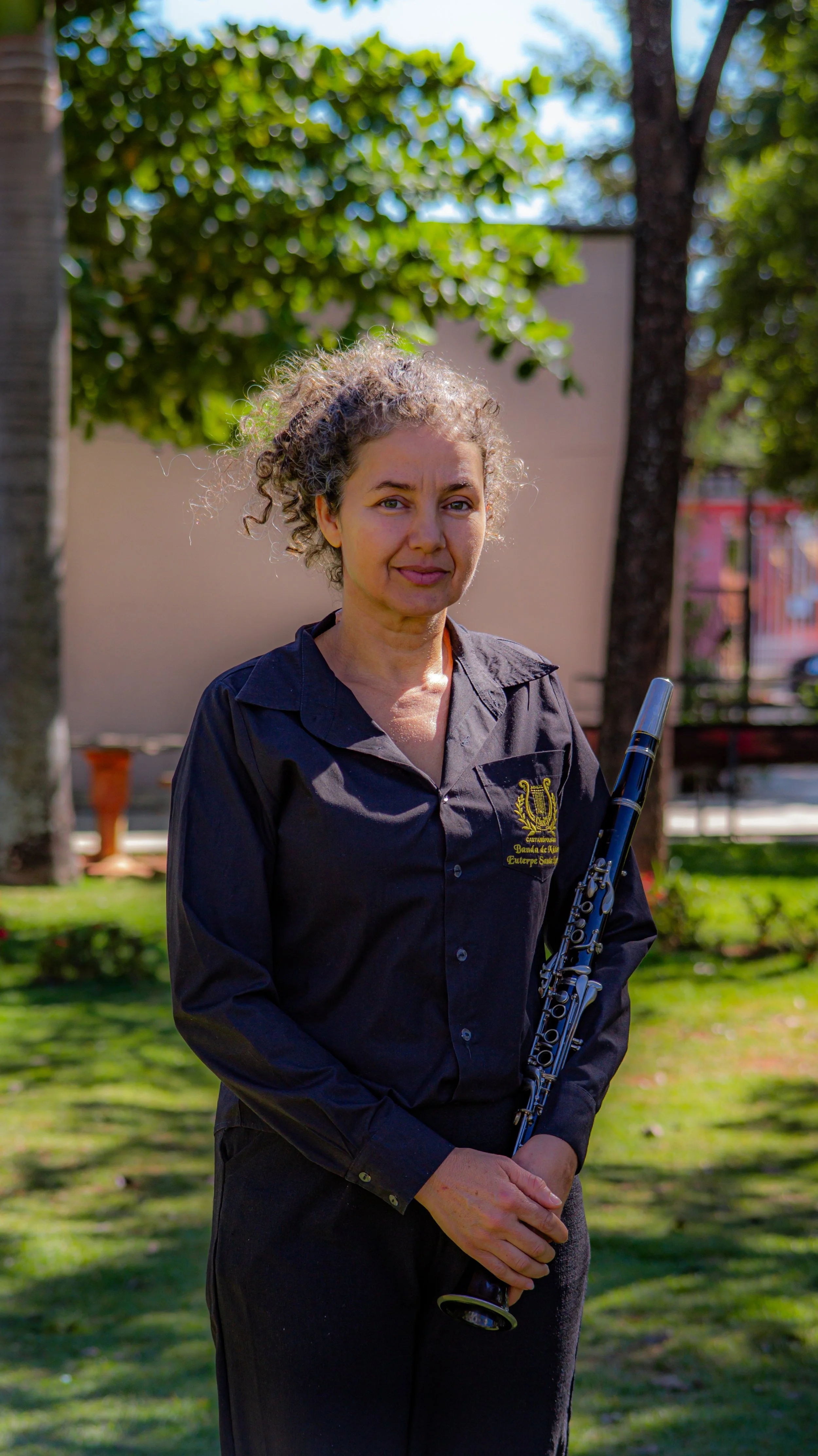 Mulher segurando uma flauta, de cabelo cacheado, vestindo uma camisa preta, ao ar livre com árvores e grama ao fundo.