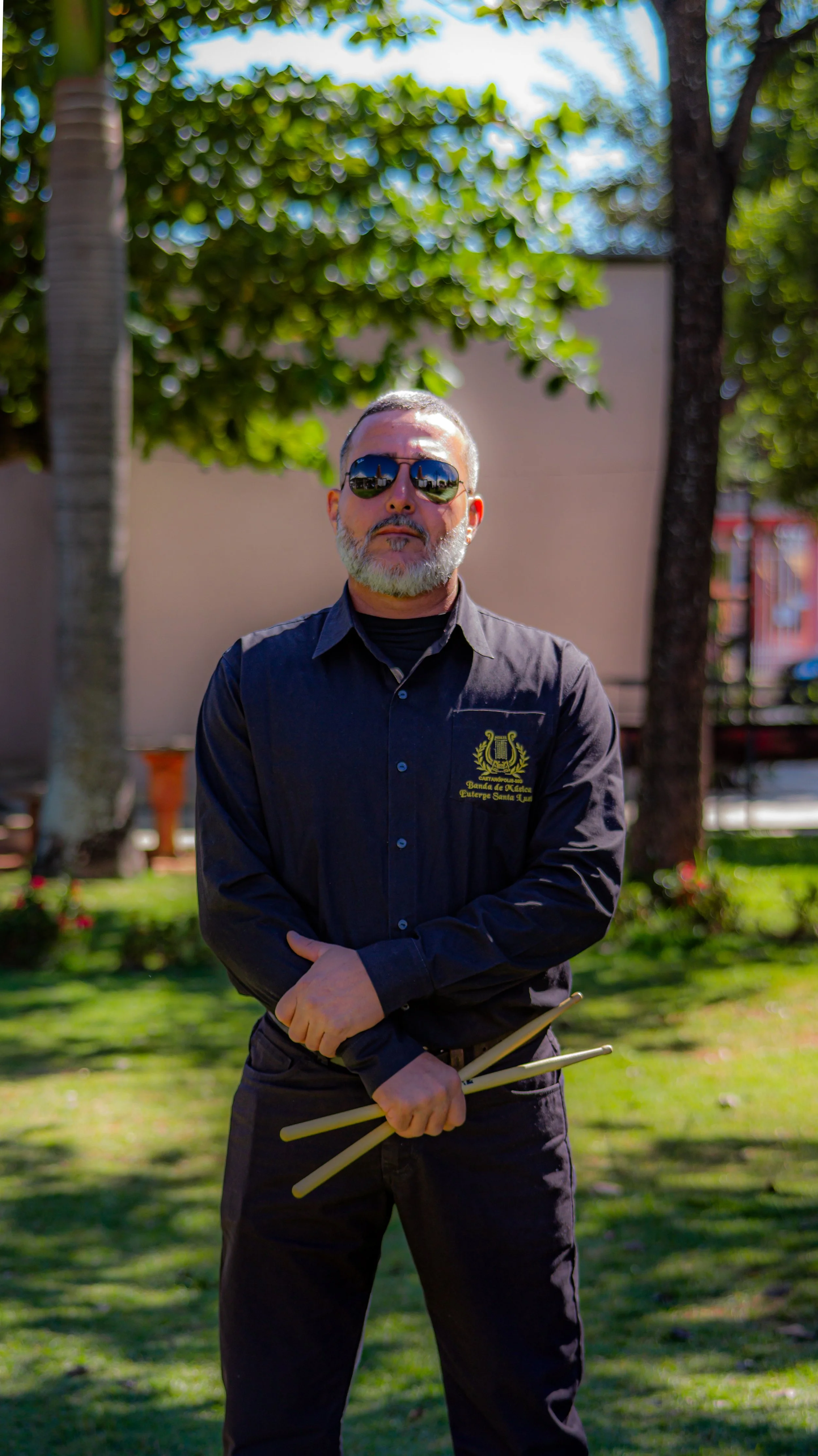 Homem com barba grisalha, chapéu preto e óculos escuros, vestindo camisa preta com logotipo e palavras em português, segurando duas baquetas, em um parque com árvores e gramado.