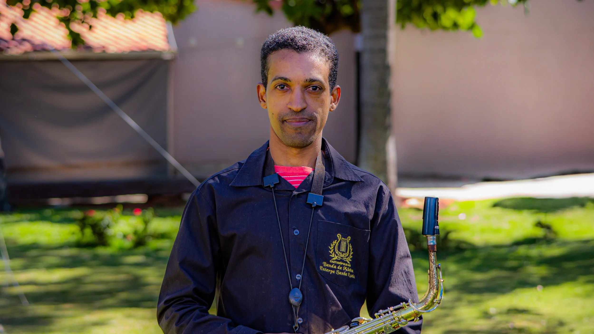 Homem segurando um saxofone ao ar livre, trajando camisa preta com brasão na manga e uma faixa vermelha com branco visível no pescoço, na frente de árvores e parede. 