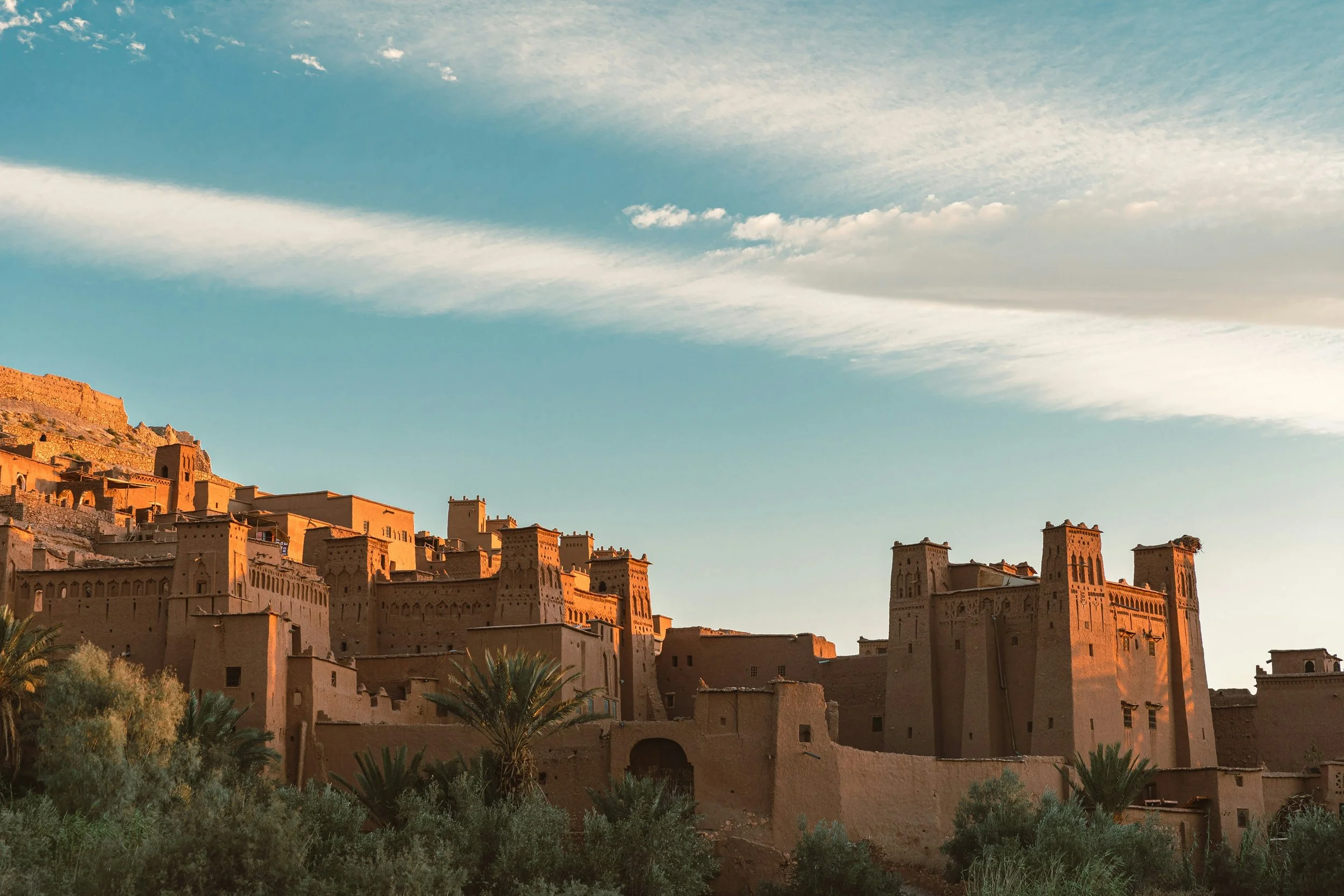 Ancient desert fortress with high walls and towers, surrounded by greenery, under a bright sky with wispy clouds.