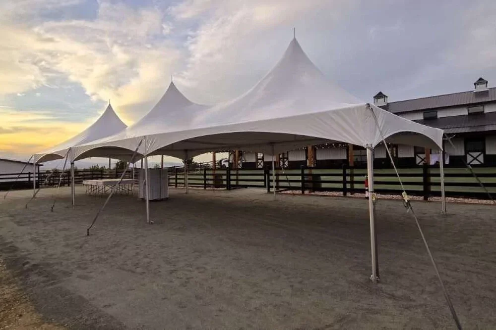 Large white event tent set up on a dirt surface with a sunset sky in the background, adjacent to a barn-like building with a fenced area.
