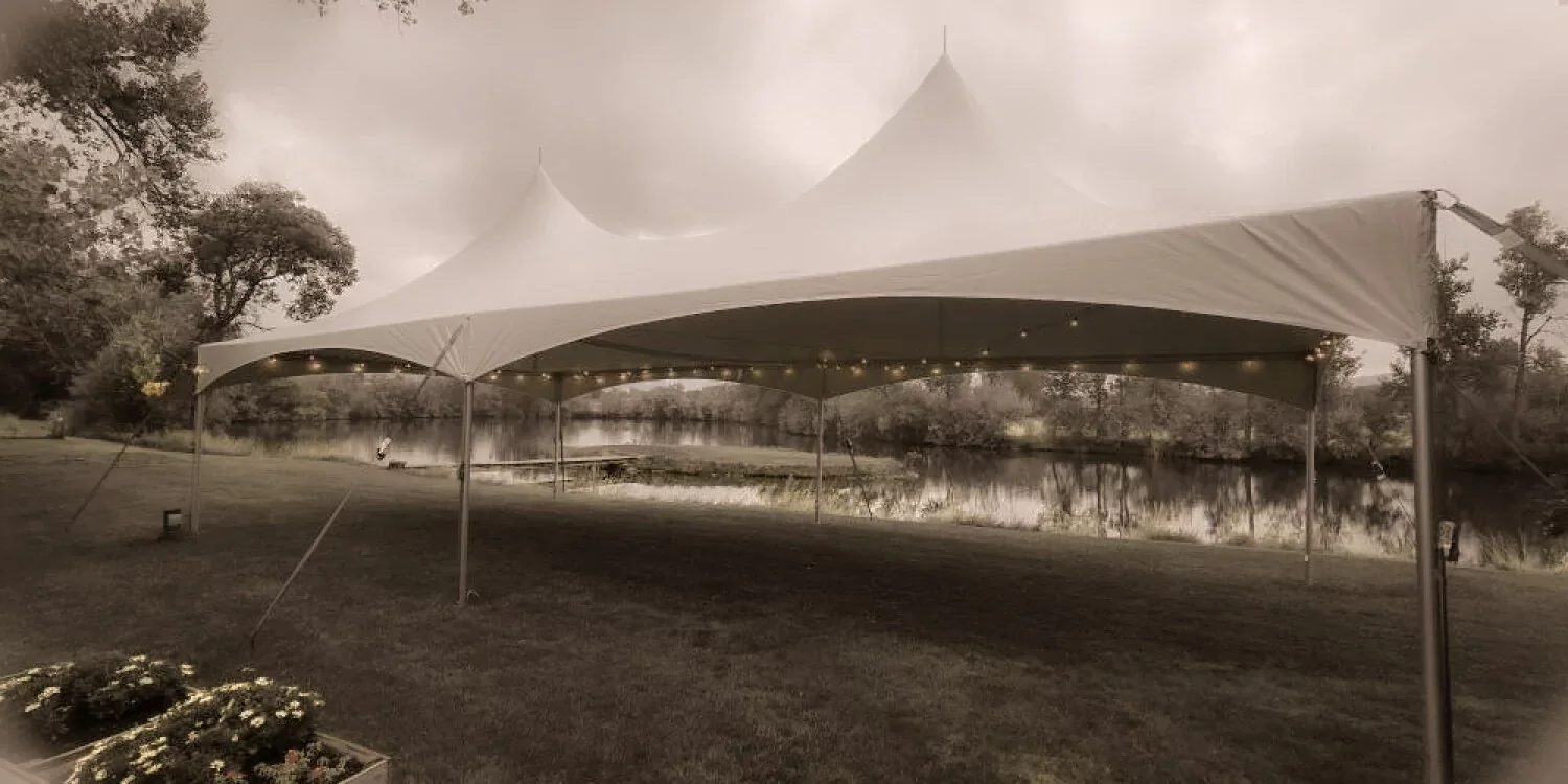 A large outdoor tent set up near a body of water, with trees in the background and string lights hanging inside the tent.