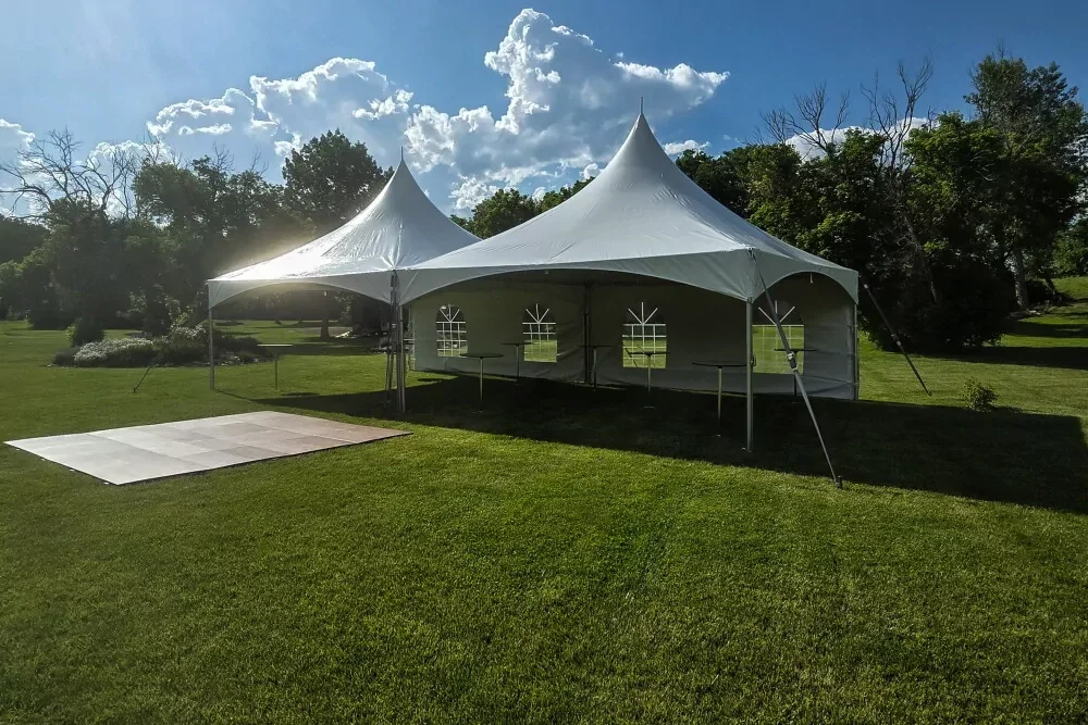 A white event tent set up on a green lawn with a small white patio in front of it, surrounded by trees and under a partly cloudy sky.